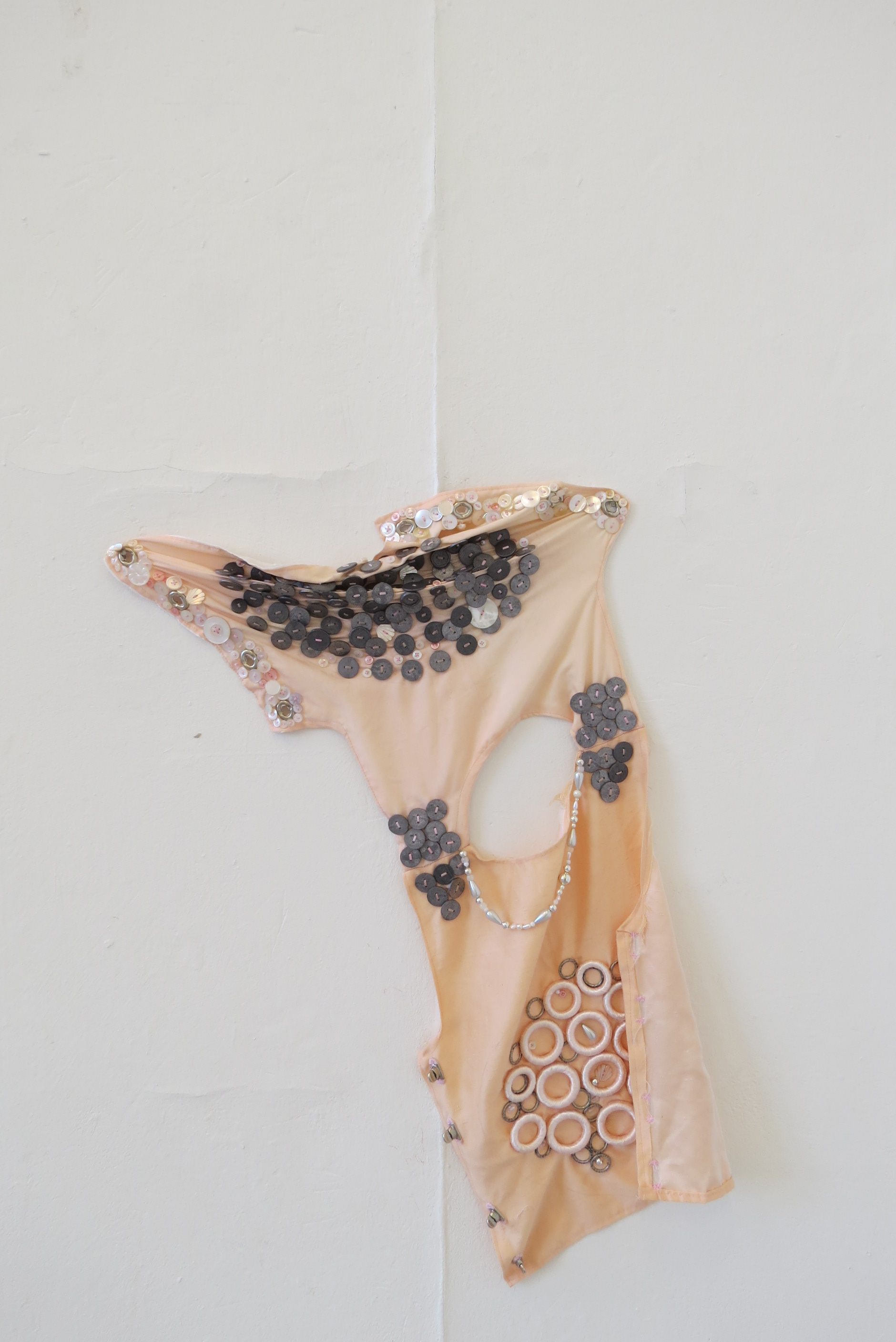 Fabric body armour, which is peach in colour and decorated using plastic and glass beads and buttons, circular metal weights and satin ribbon covered wooden rings, is hung on the wall.