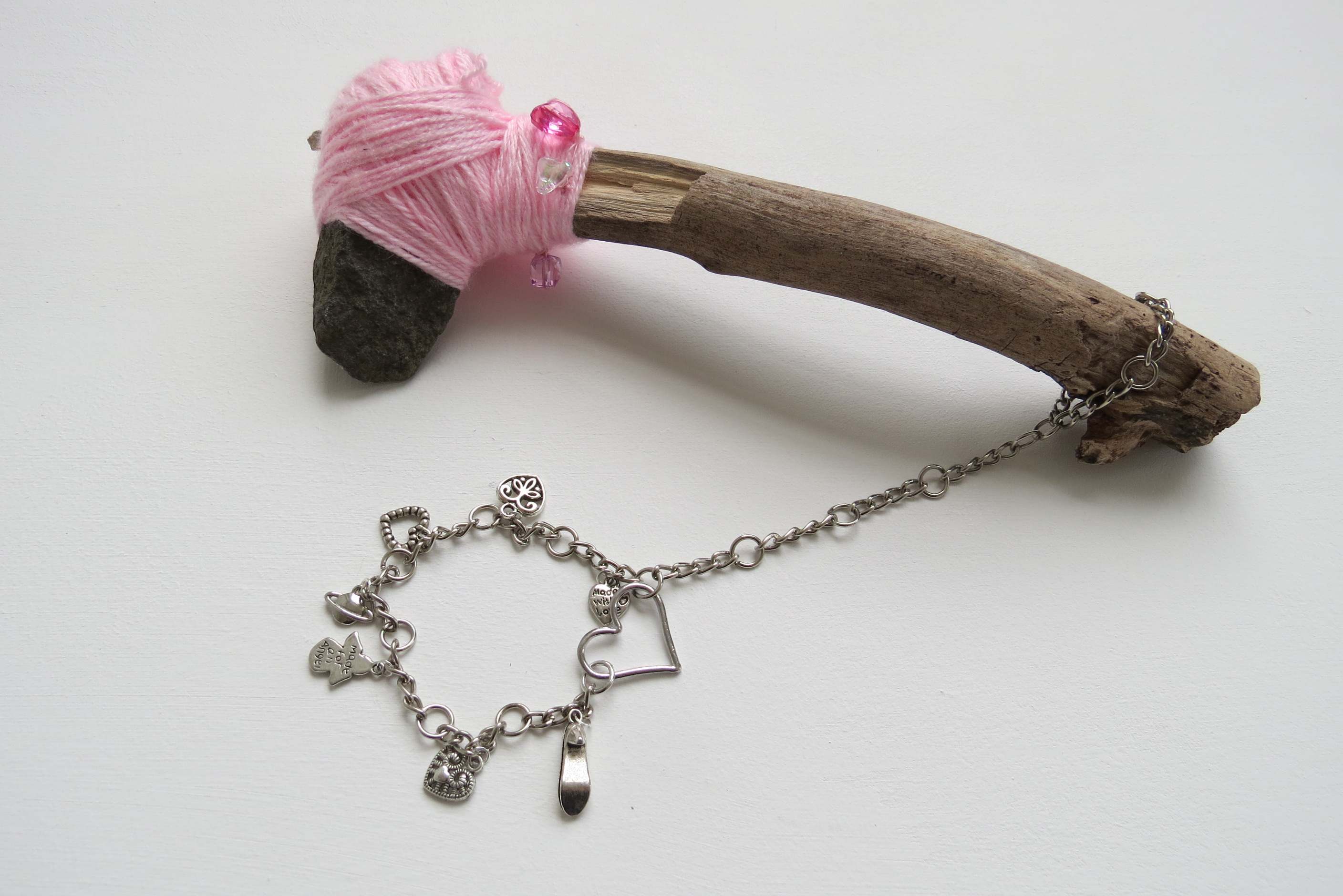 An axe created using a stone bound by pink yarn to a stick. Glass and plastic beads adorn the yarn, with a metal chain wrist-strap with metal charms attached.