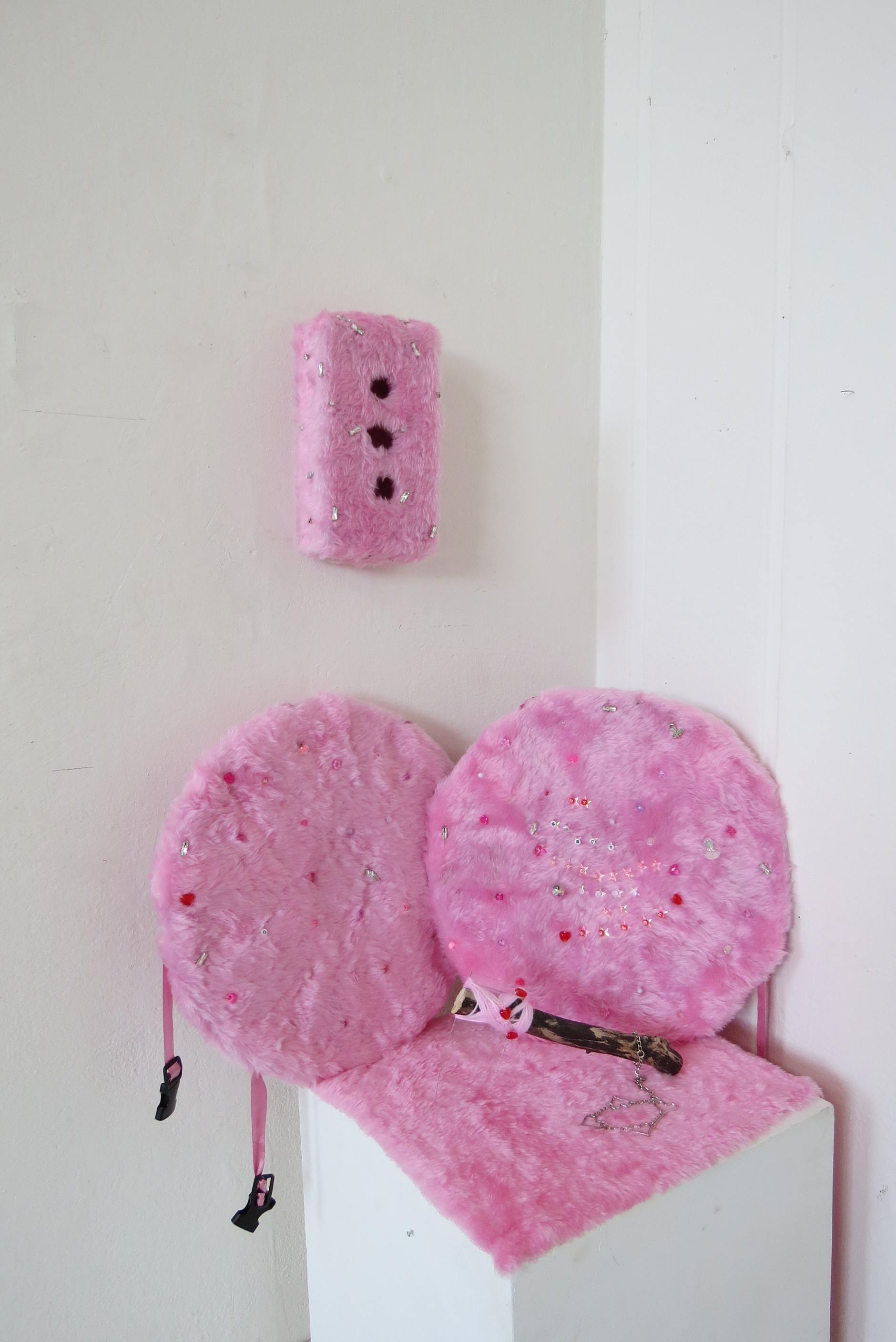 A brick made of fuzzy pink fabric with the same fabric covering body armour shields from car hub caps and an axe made from a shard of glass, bound to a stick with pink yarn.