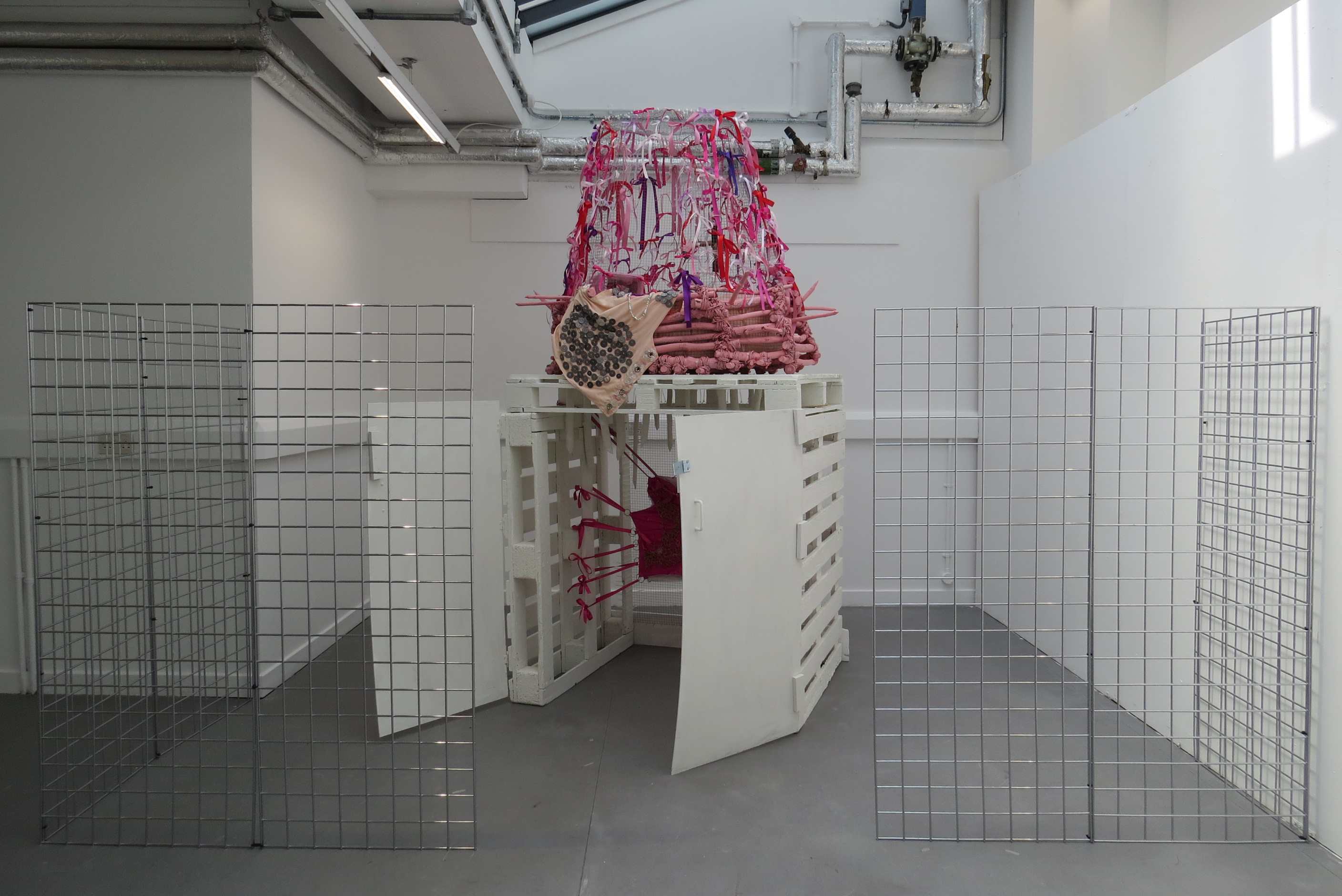 A large structure made from pallets, sticks bound together to form a nest and galvanised wire to create a tent structure above. The structure is adorned with objects, ribbons, beads and buttons.