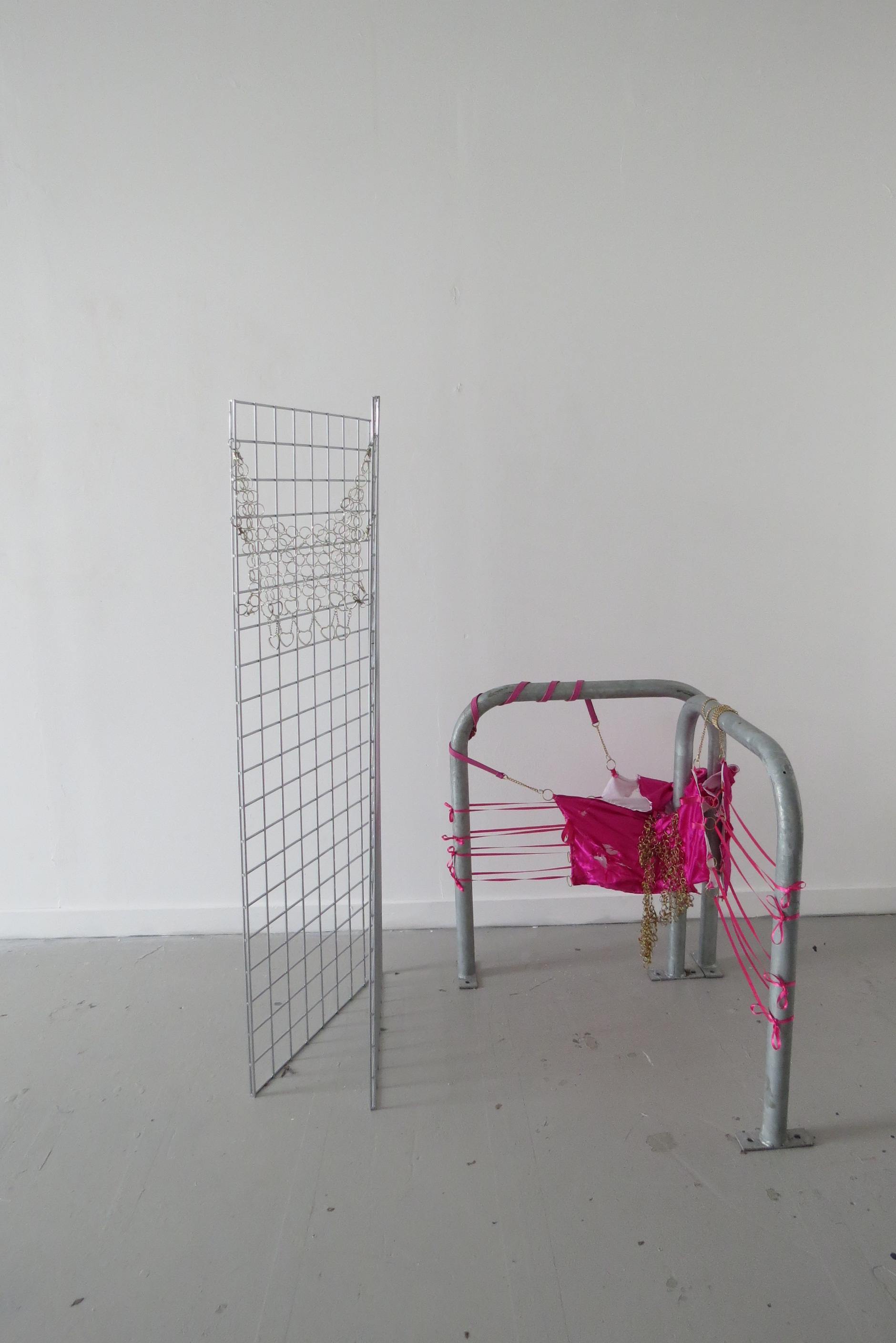 Metal gates are used to create an angular structure, with metal chain mail created using discarded key chain metal ware is hung between the gates. A fabric body armour, which is dark pink in colour, decorated with glass and plastic beads, satin ribbon covering wooden rings and gold metal ware, is hung from two bicycle stands. 