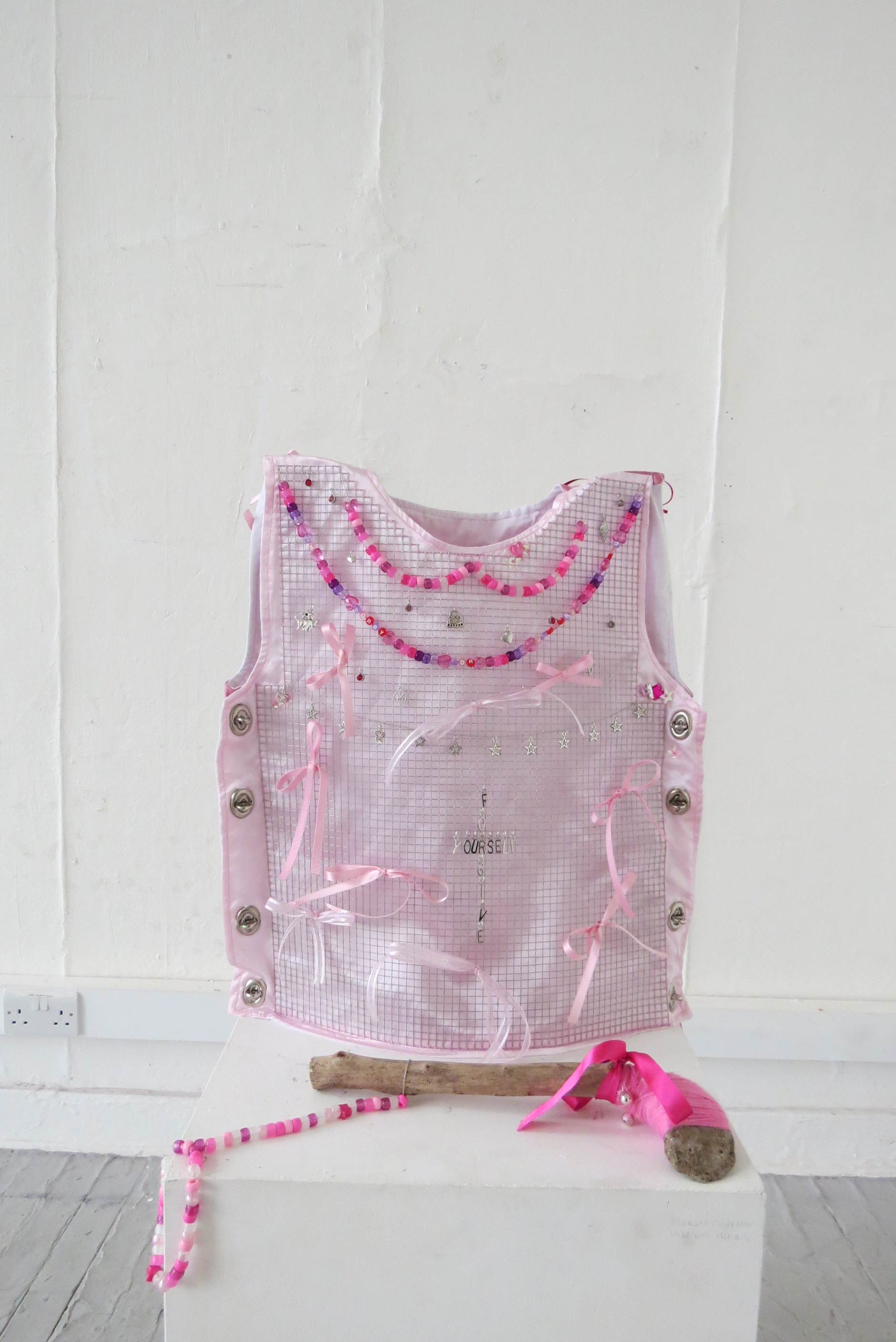 A fabric body armour with galvanised wire spread across the chest, decorated with glass and plastic beads and charms, a weapon made from a stone bound to a stick with pink yarn placed in front.