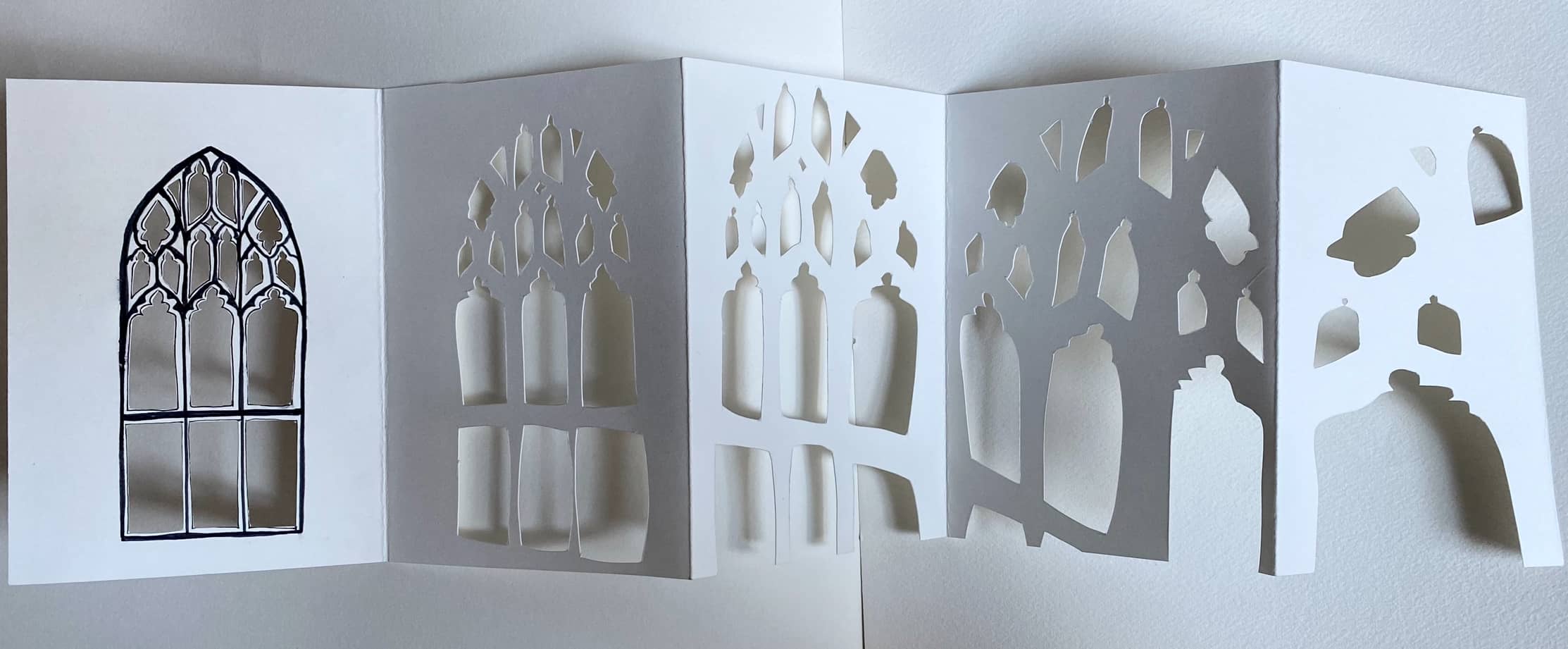 a stretched white paper concertina book showing 6 panels of cut out shapes, starting with a regular cu=hurch window outlined in black ink, then ending with an abstracted and distorted jumble of shapes