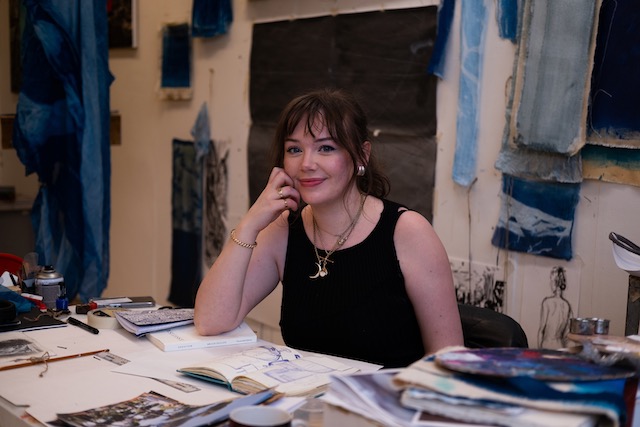 Person at studio desk, messy papers and draping fabrics in various blue tones in the background