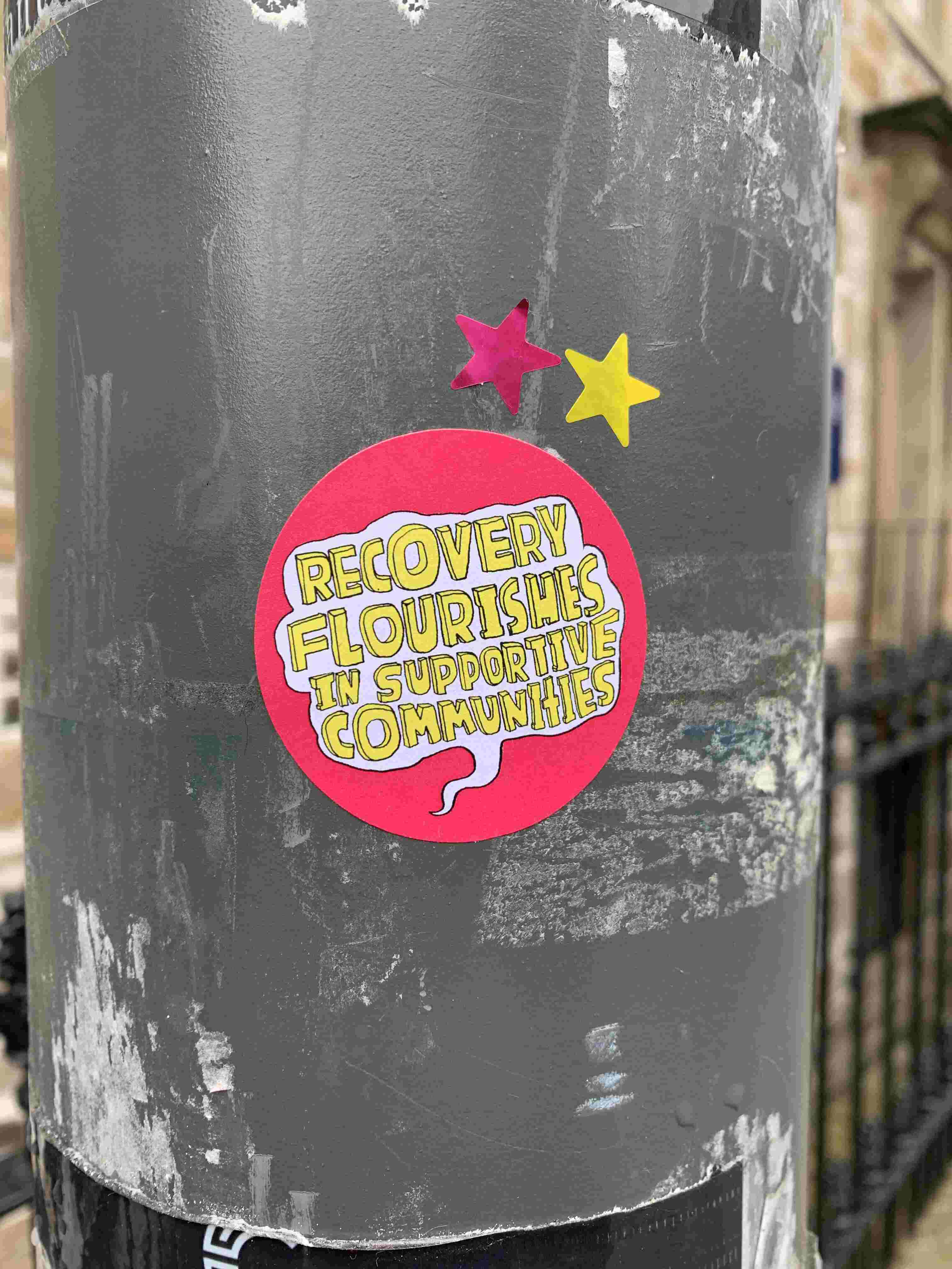 Pink sticker on lampost with white speech bubble and yellow text saying: "recovery flourishes in supportive communities".