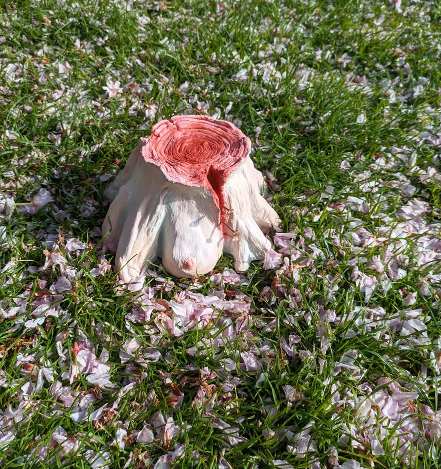 A tree stump made of human flesh, sat atop grass and flower petals outside.