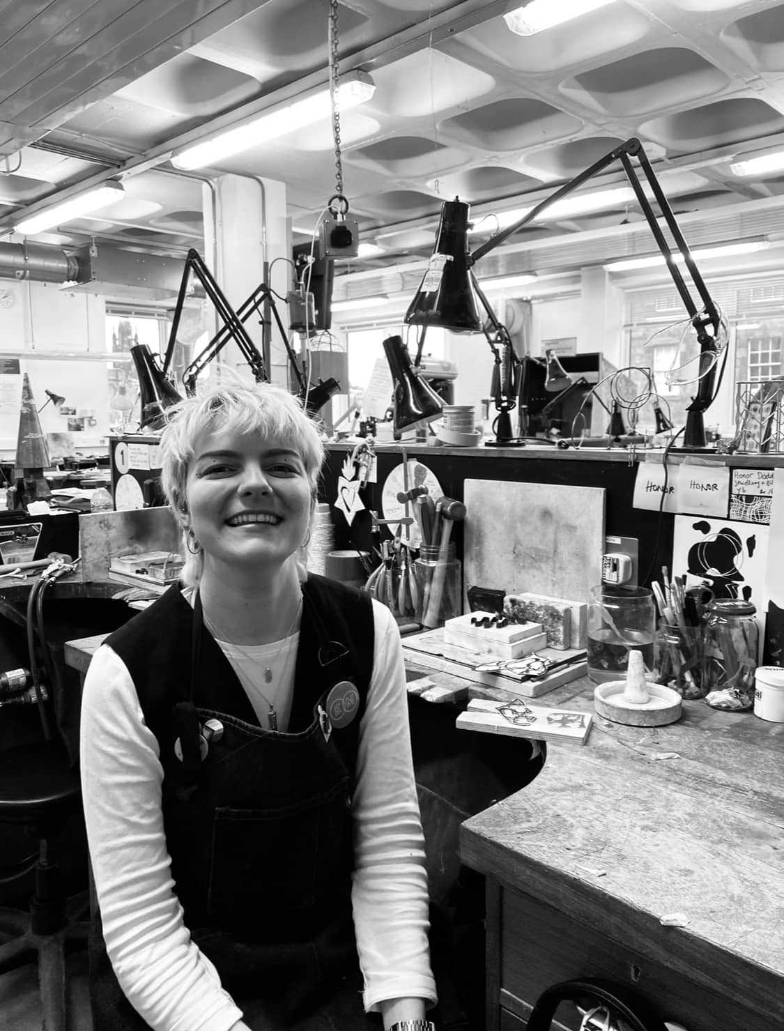 a young white person with bleached hair and wearing an apron sits at a jewellery bench facing the camera with a smile on her face. the photo is black and white