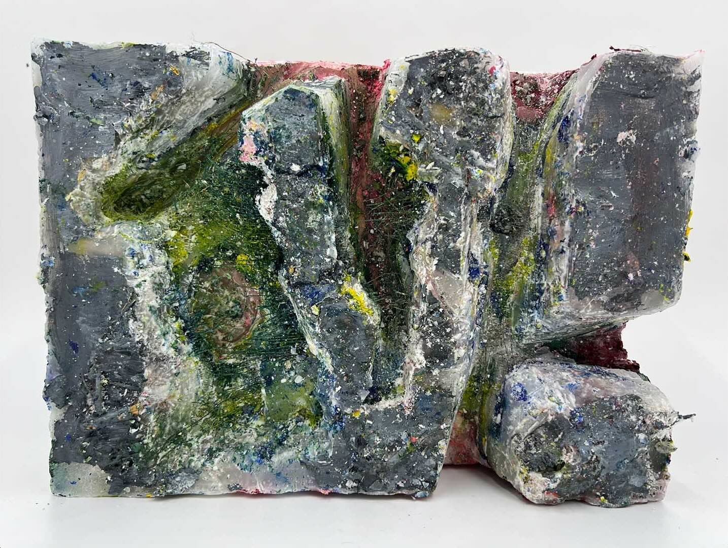 photograph of block of wax and paint that has been carved into. it is grey on the non-carved places, but the carved places show different paint colours, frayed fabric, and layers of wax.