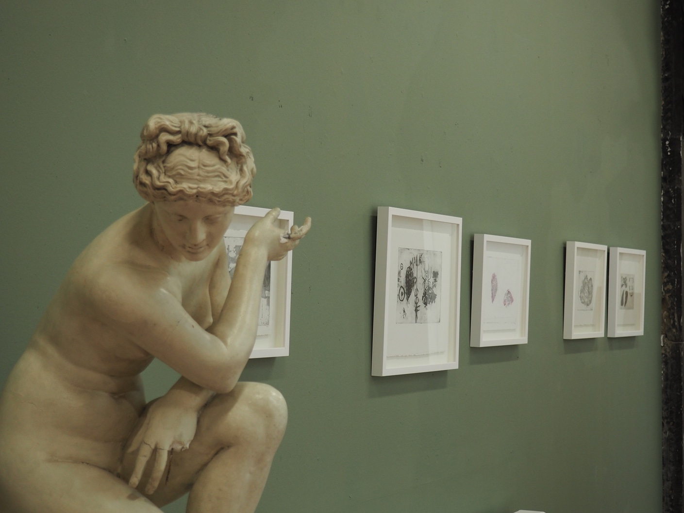 Plaster cast of Venus with damaged hands, gesturing towards framed prints against a green wall