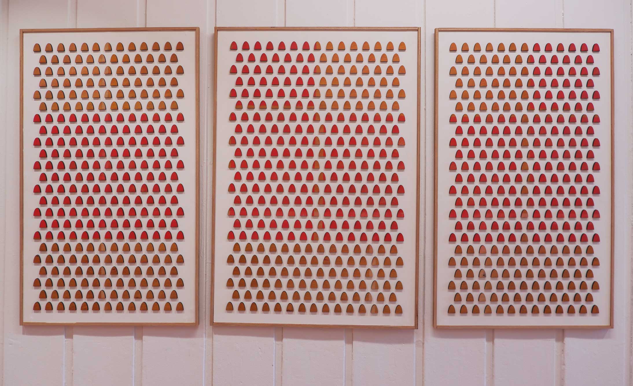 Three framed white boards each with a grid of wooden gravestone shaped pieces of wood. Some of the wooden pieces are sprayed red in an abstract pattern.