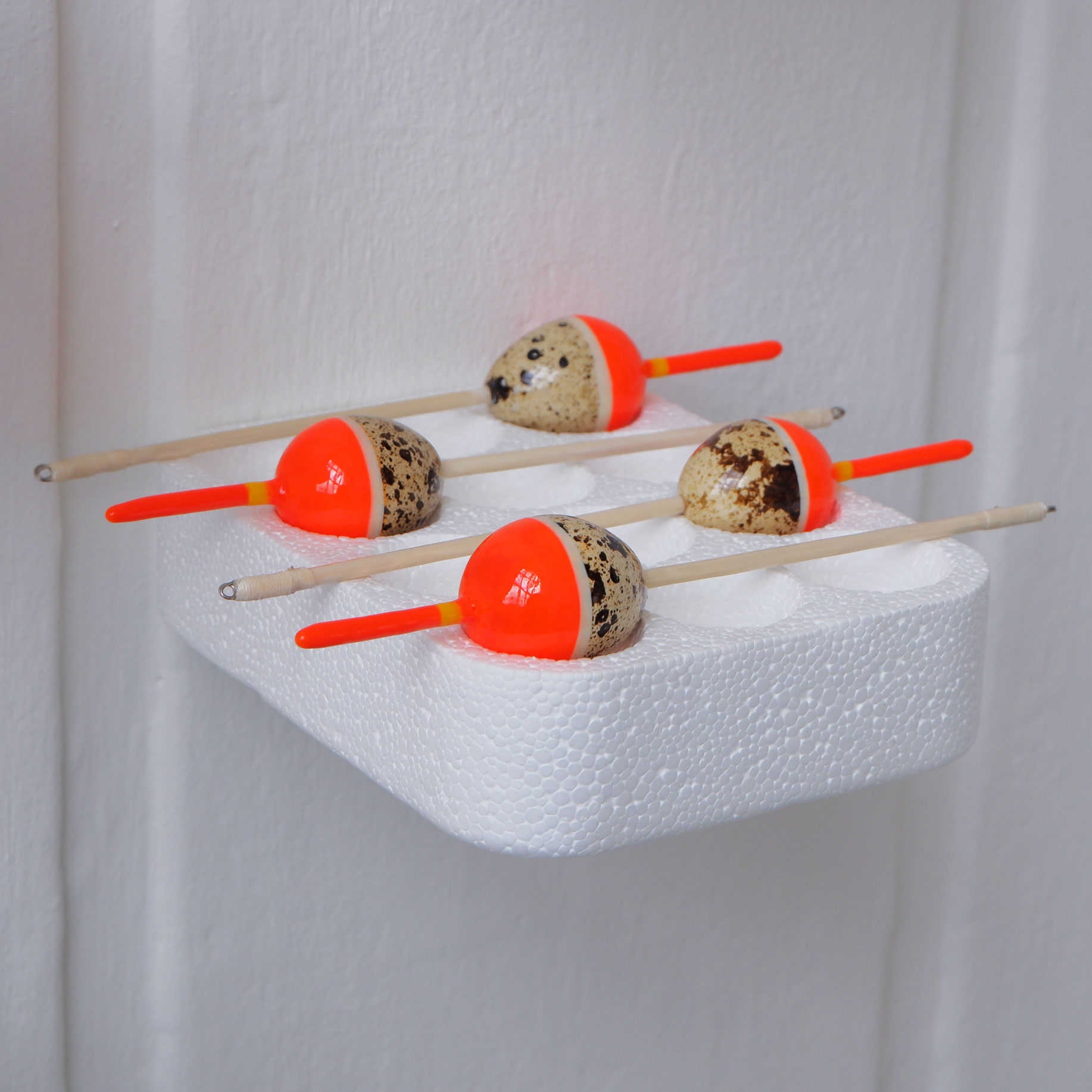 Four quail egg bobbers resting on a polystyrene tray attached to a white wall.