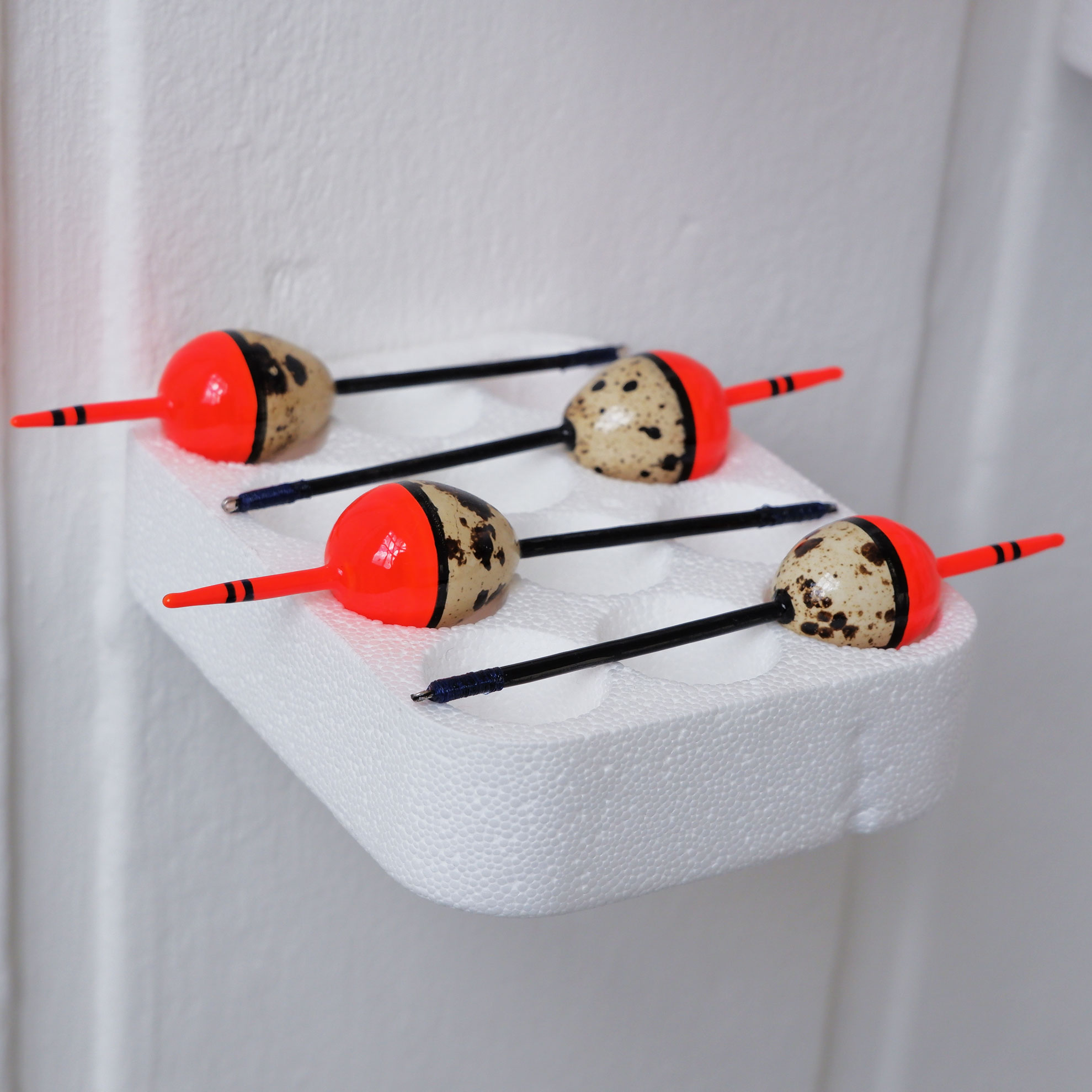 Four quail egg bobbers resting on a polystyrene tray attached to a white wall.