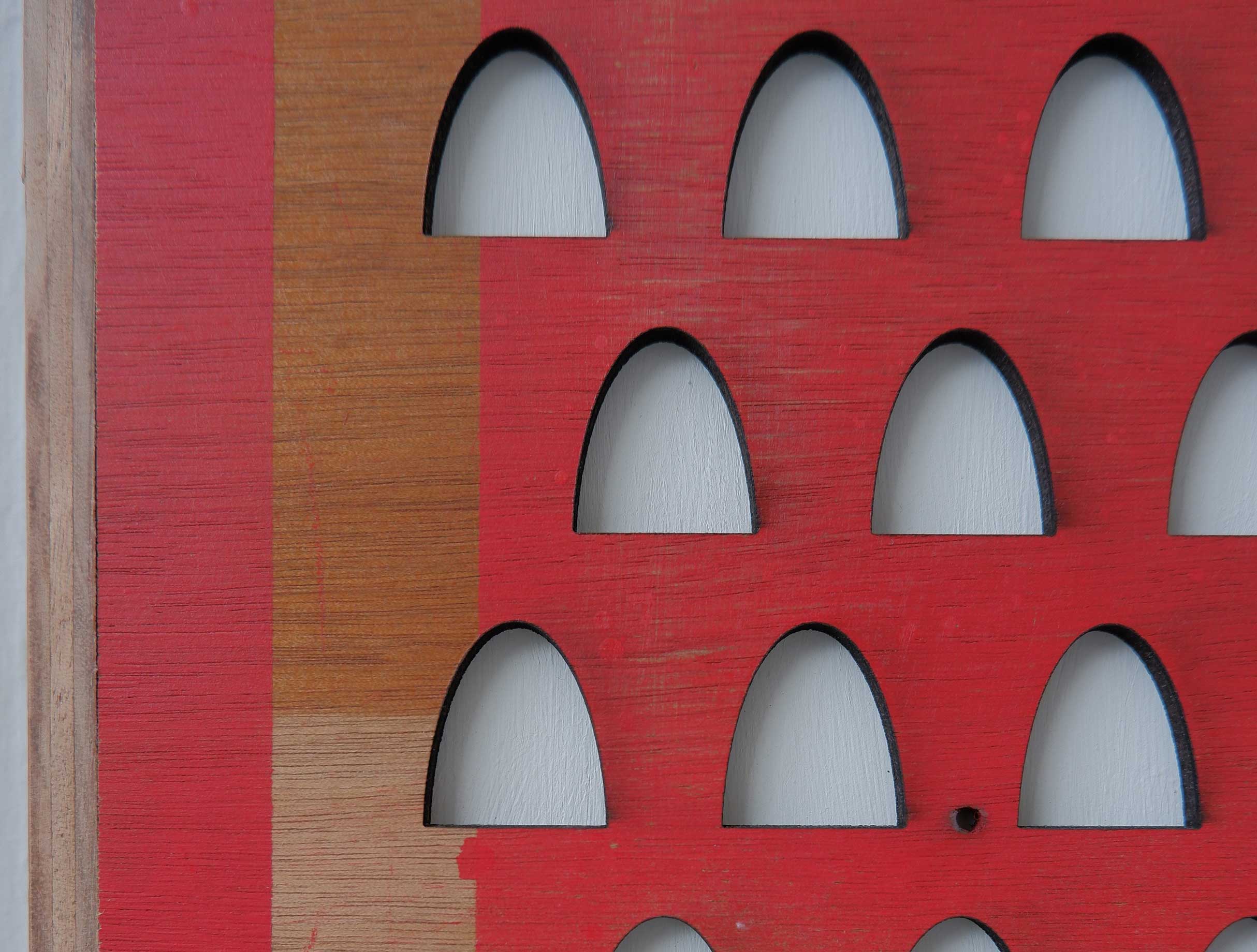 A detail image of a framed board out of which dovecot/gravestone shapes have been removed to reveal the white board underneath. Some of the board is sprayed red in an abstract pattern.