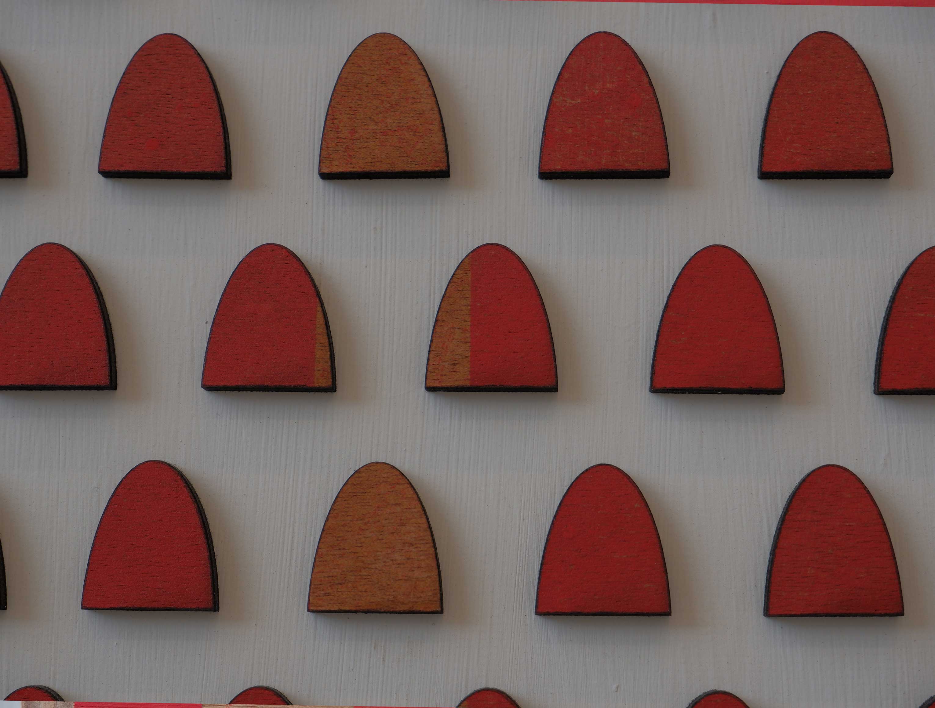 A white board with a grid of wooden gravestone shaped pieces. Some of the wooden pieces are sprayed red in an abstract pattern.