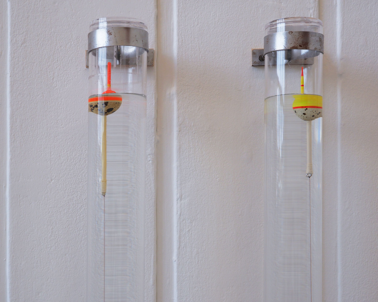 Detail image of two clear acrylic tubes mounted on a white wall, attached by a steel bracket. Each tube contains water in which floats one quail egg bobber attached to red fishing line