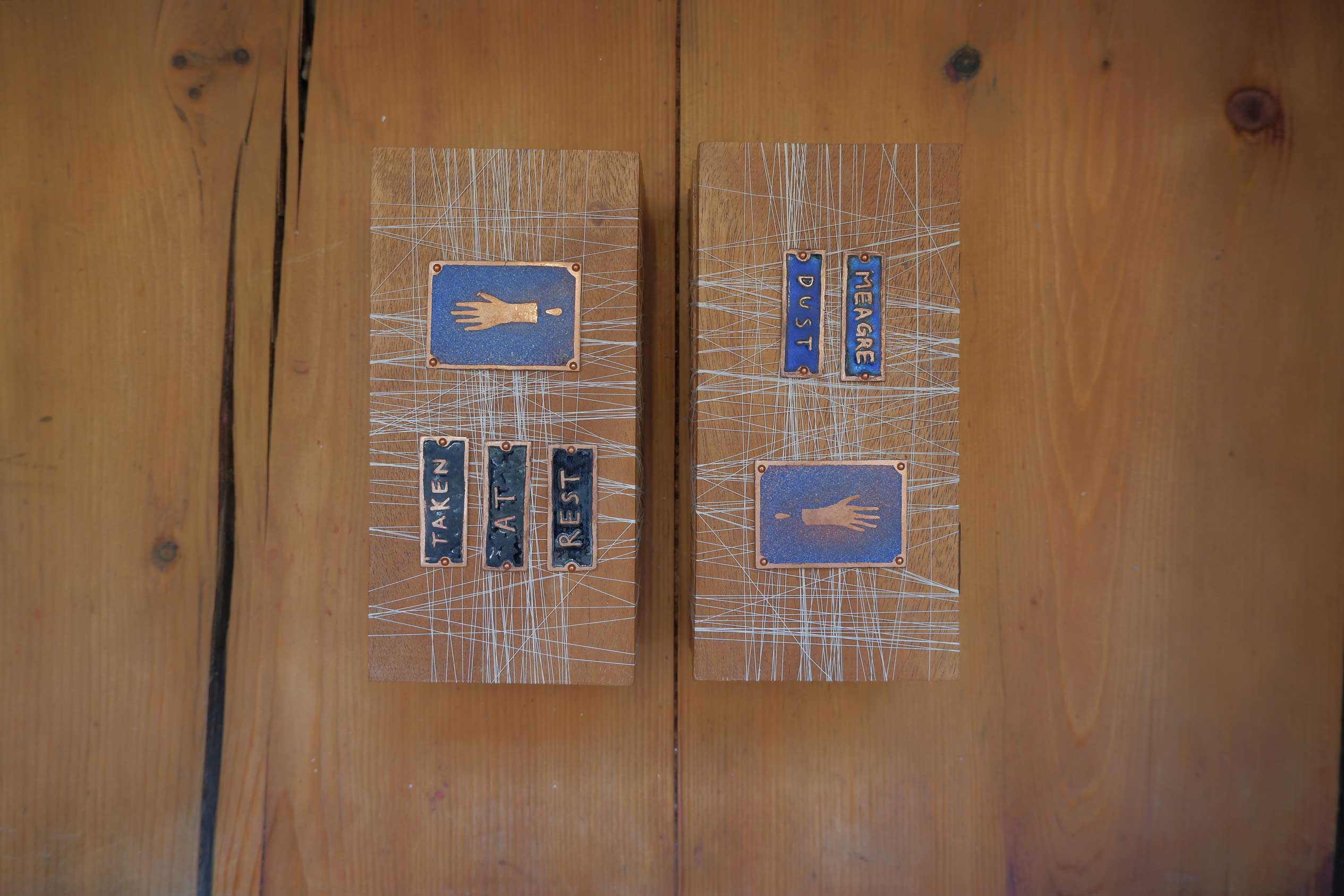 Two wooden boxes laid next to each other, they are decorated with crisscrossed white thread and blue enamel tiles. The tiles on one box display the poem 'Meagre Dust' and those on the other show the poem 'Taken At Rest'. Both boxes each have one enamel tile with the image of a bleeding hand on them.