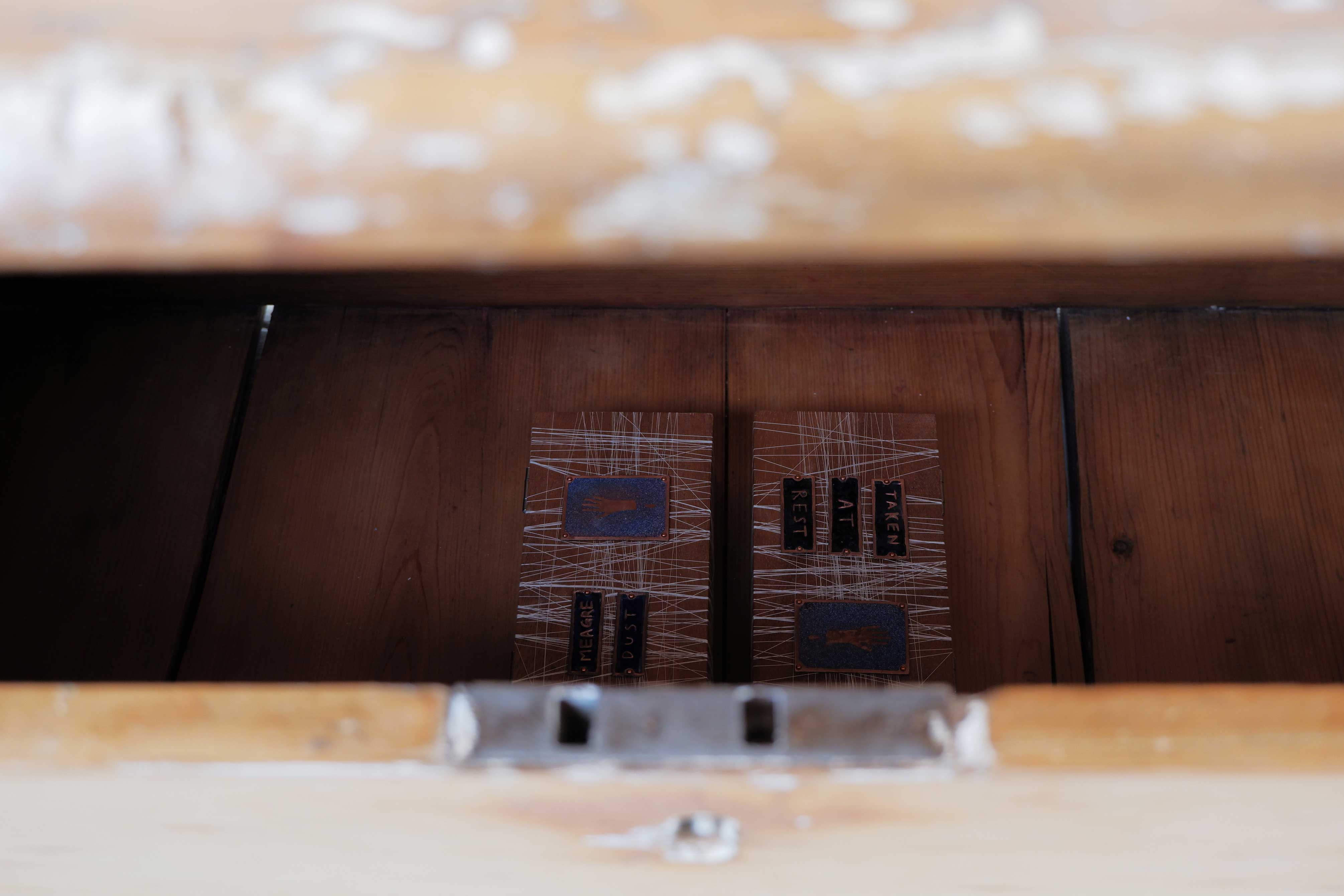 Two wooden boxes laid next to each other, they are decorated with crisscrossed white thread and blue enamel tiles. The tiles on one box display the poem 'Meagre Dust' and those on the other show the poem 'Taken At Rest'. Both boxes each have one enamel tile with the image of a bleeding hand on them