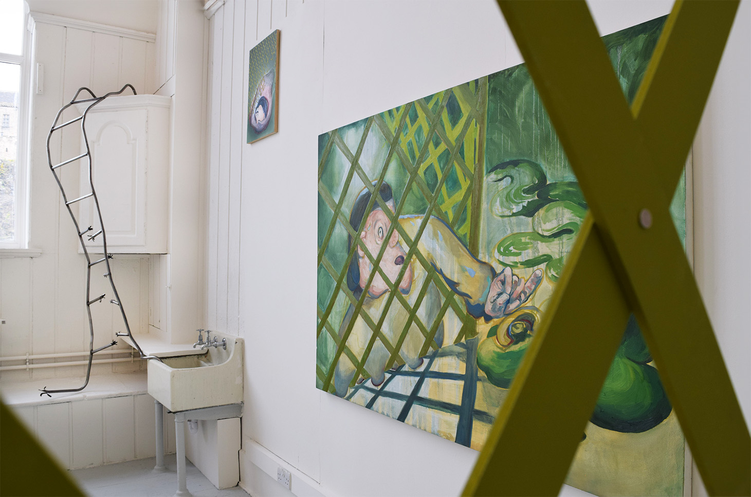 Image is of a steel ladder that snakes into the sink, two green paintings of blobby figures that reach through a green fence and a green lattice fence in the forefront of the image which as a viewer we are looking through.
