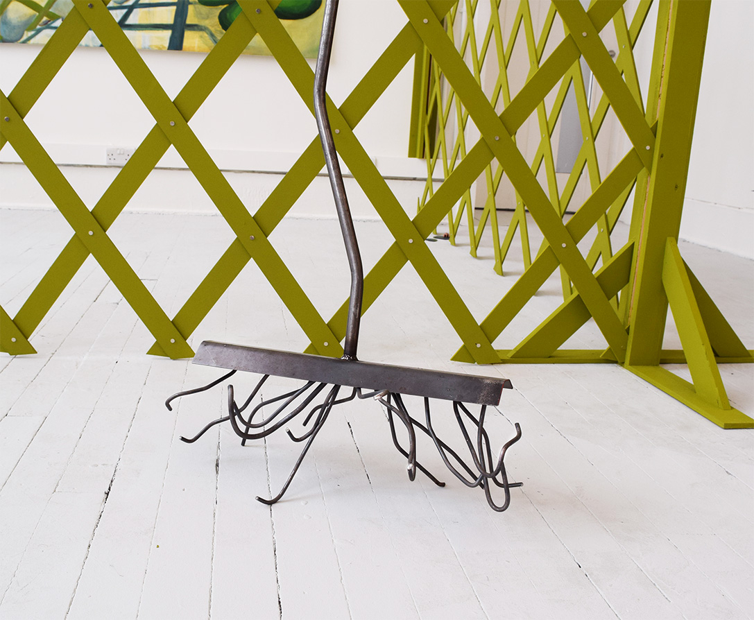 Image is a close up of a self-standing steel rake sculpture that has unruly spikes. In the background is the green lattice fence. 