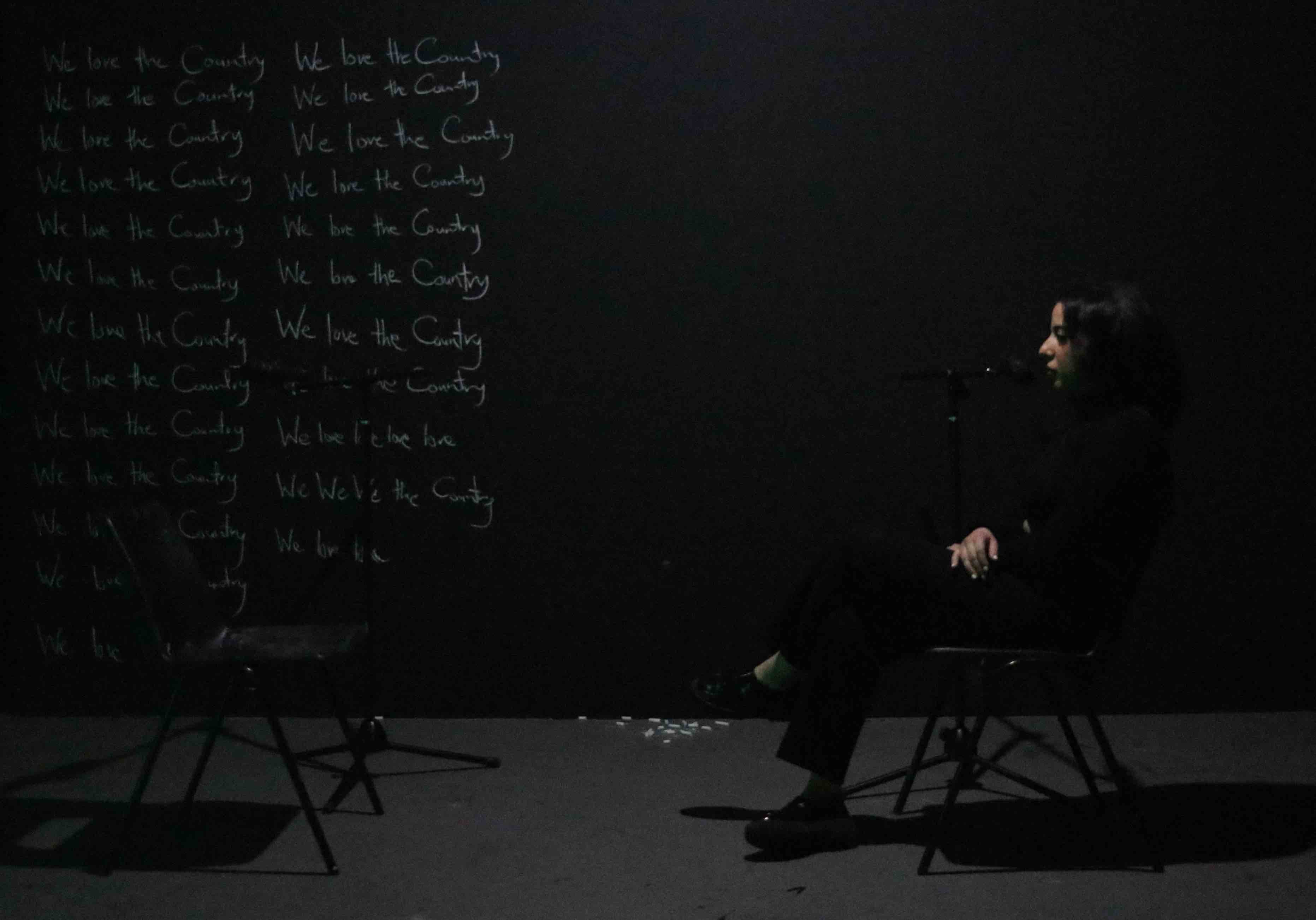 A person dressed in black sits on a chair facing another empty chair in a dark room. They are speaking into a microphone. The wall behind them is black and covered with the repeated handwritten phrase "We love the Country" in white chalk. The room is dimly lit, casting shadows on the floor.