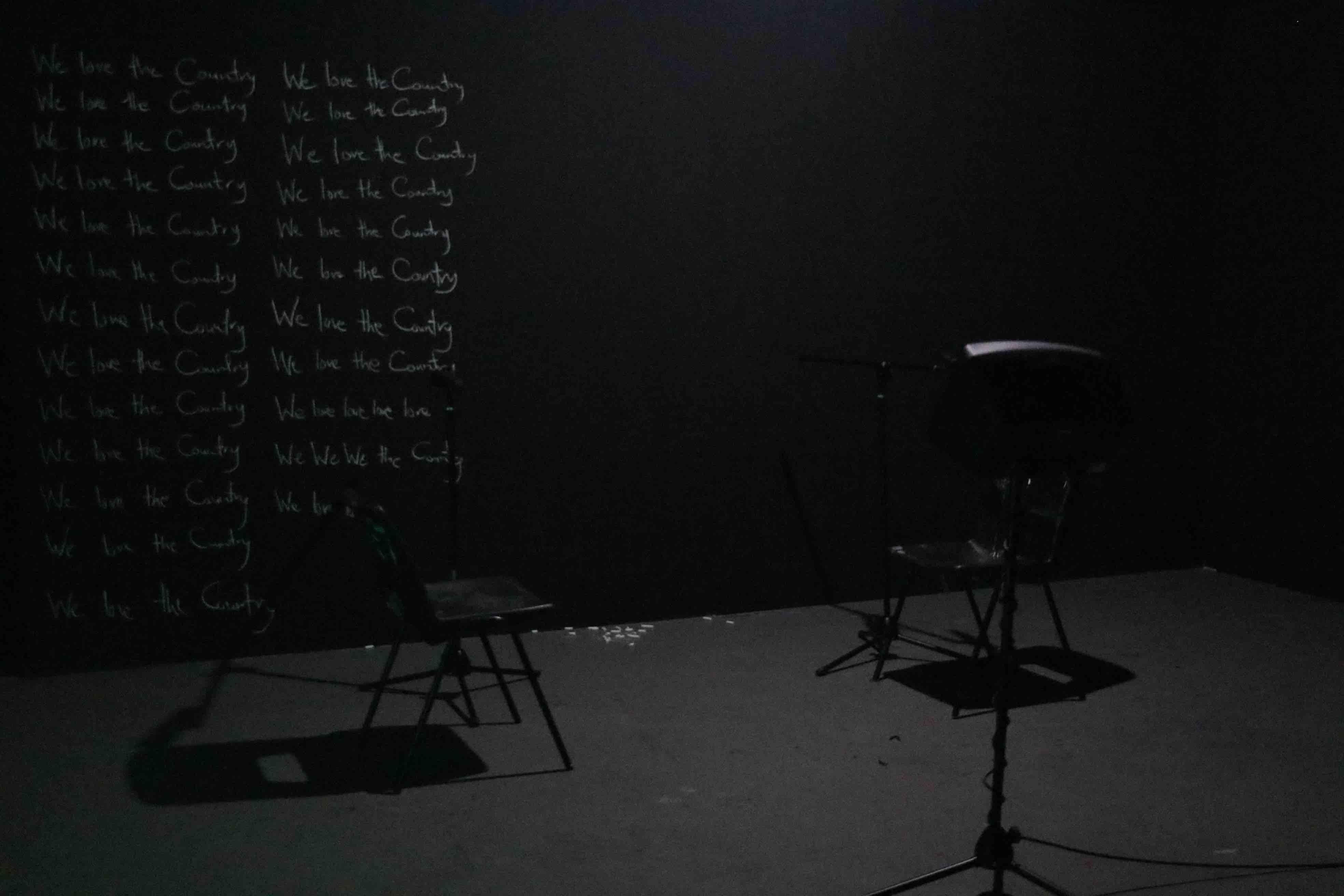 two stools in front of a black wall covered with the repeated phrase "We love the Country" written in white chalk.  between the stools is a speaker. The background is dark, and the room is lightly dimmed. 