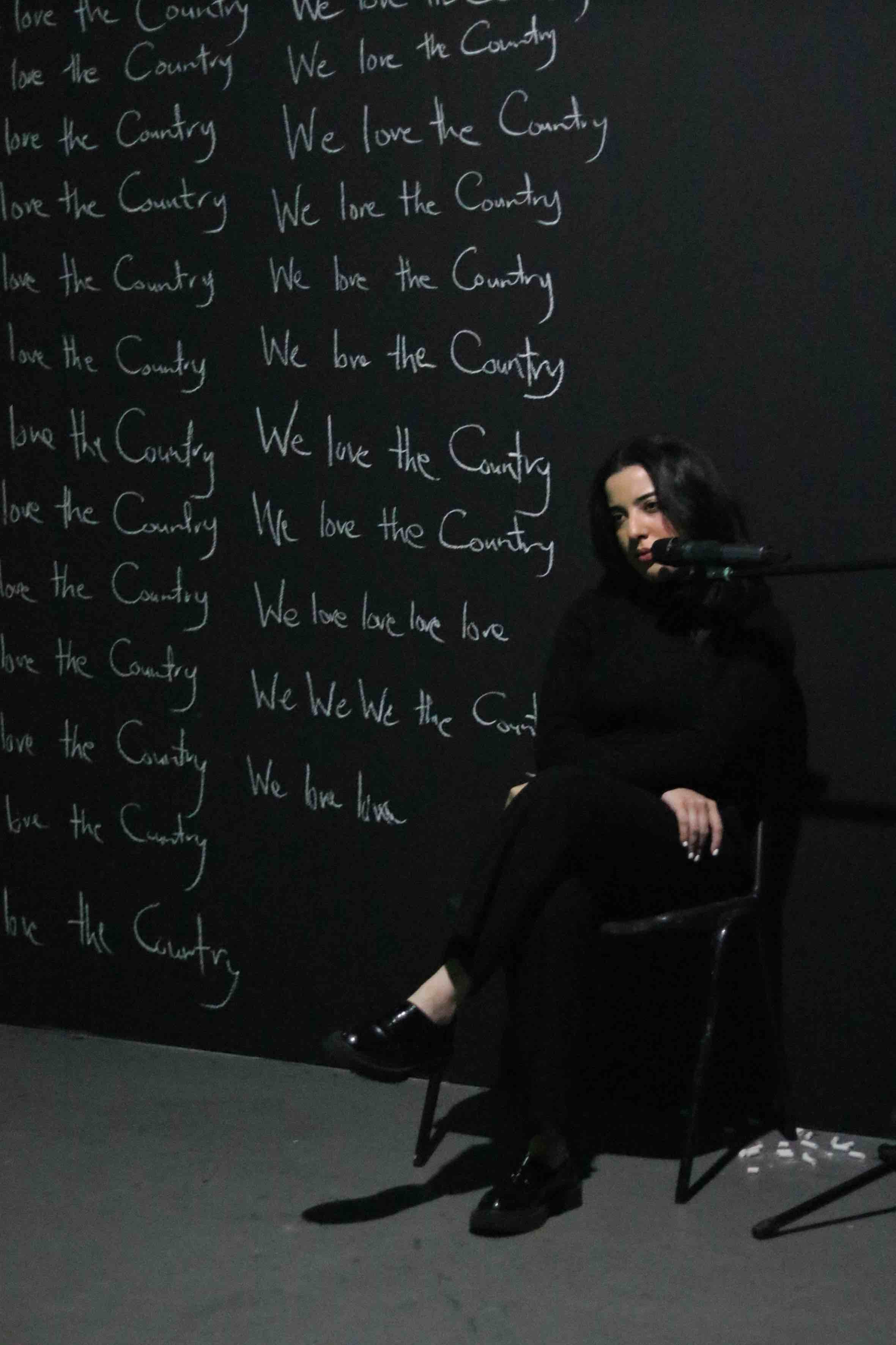 A woman dressed in black sits on a stool in front of a black wall covered with the repeated phrase "We love the Country" written in white chalk. She has long dark hair and is positioned next to a microphone stand. The background is dark, emphasizing the repetitive text and the woman's solemn expression.