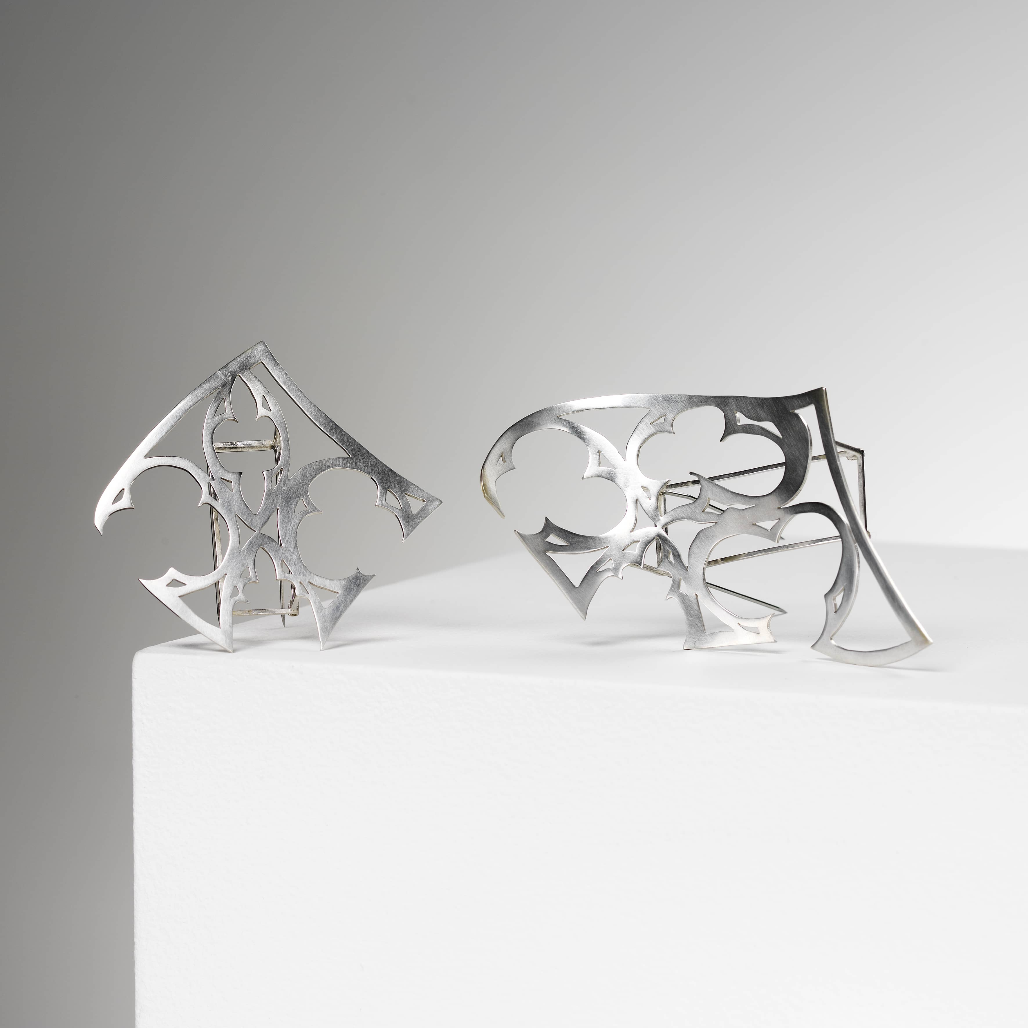 2 brooches sat side by side. the first is a diamond shaped silver cut out of a section of a gothic church window. the second is the same, but slightly stretched out and wavy. both are brooches that have a silver frame so they sit away from the body