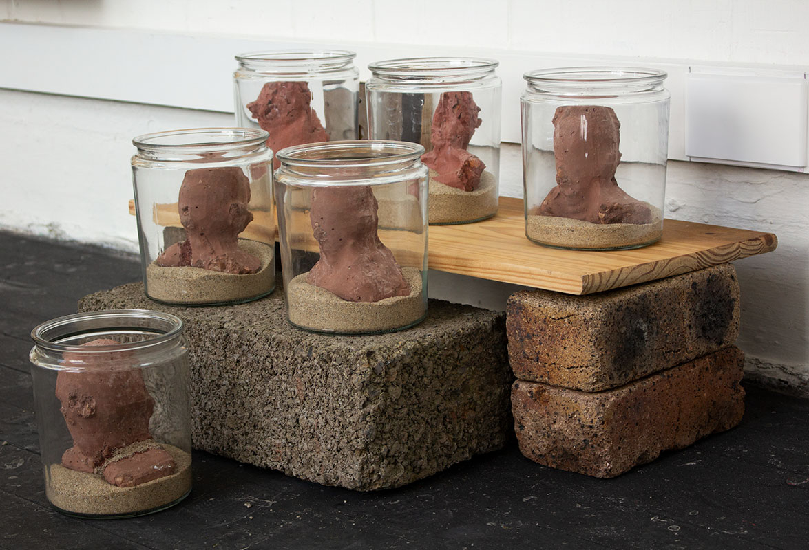 A series of concrete heads in glass jars. The Glass jars have been filled with sand and placed upon a series of bricolage plinths.