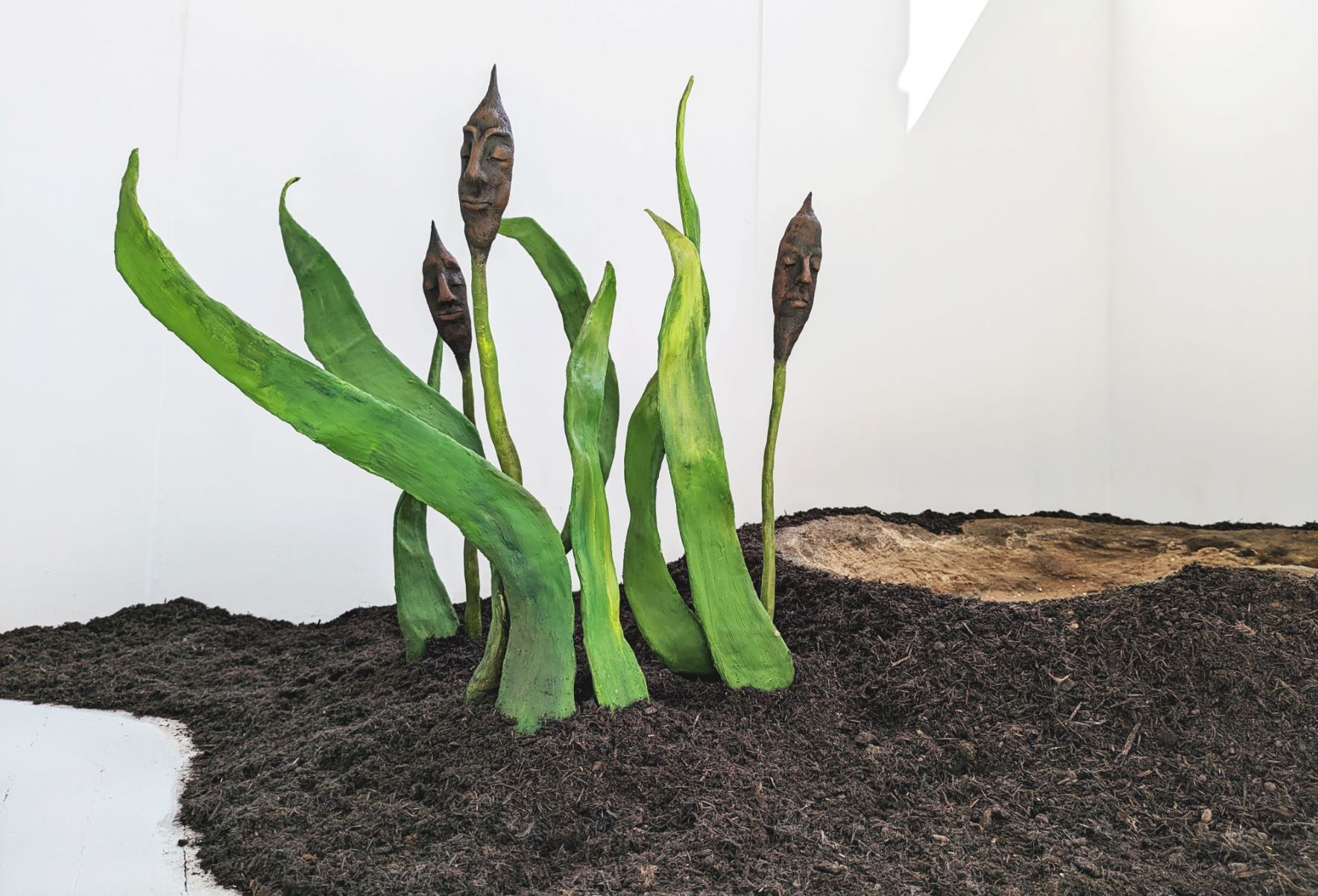 A trio of bullrushes with human faces by the edge of a pond.
