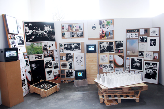 A series of boards, pedestals, and crates, each displaying seemingly disperate images. These images vary from high contast black and white drawings of orbs gritting teeth, to colourful photography, to detailed diagrams.