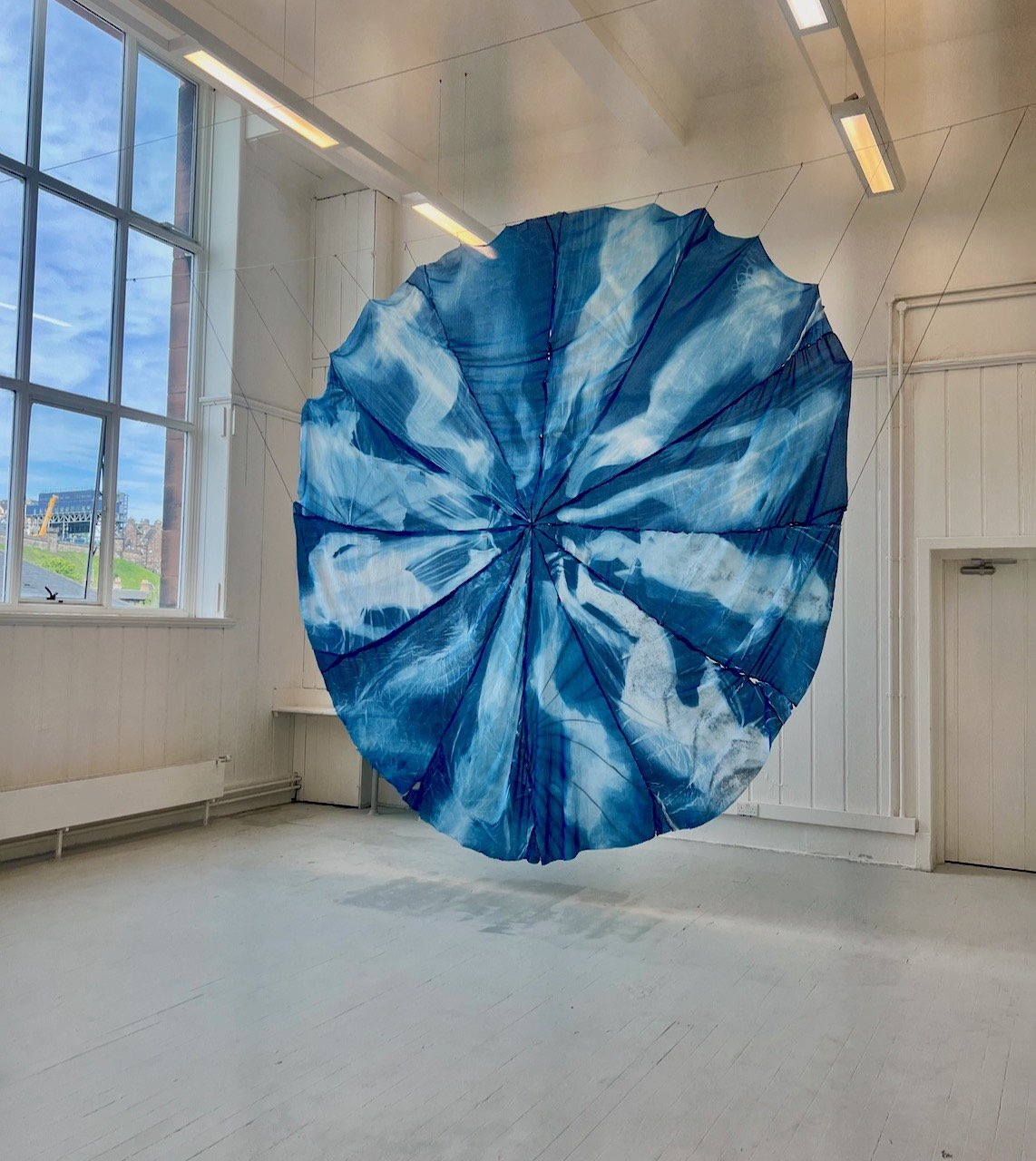 A large blue circle of fabric suspended in the air. There are 12 silhouetted figures within the circle positioned as housegments of a clock. The material is translucent and light travels through it whilst the shapes of the room can be seen from behind the work.