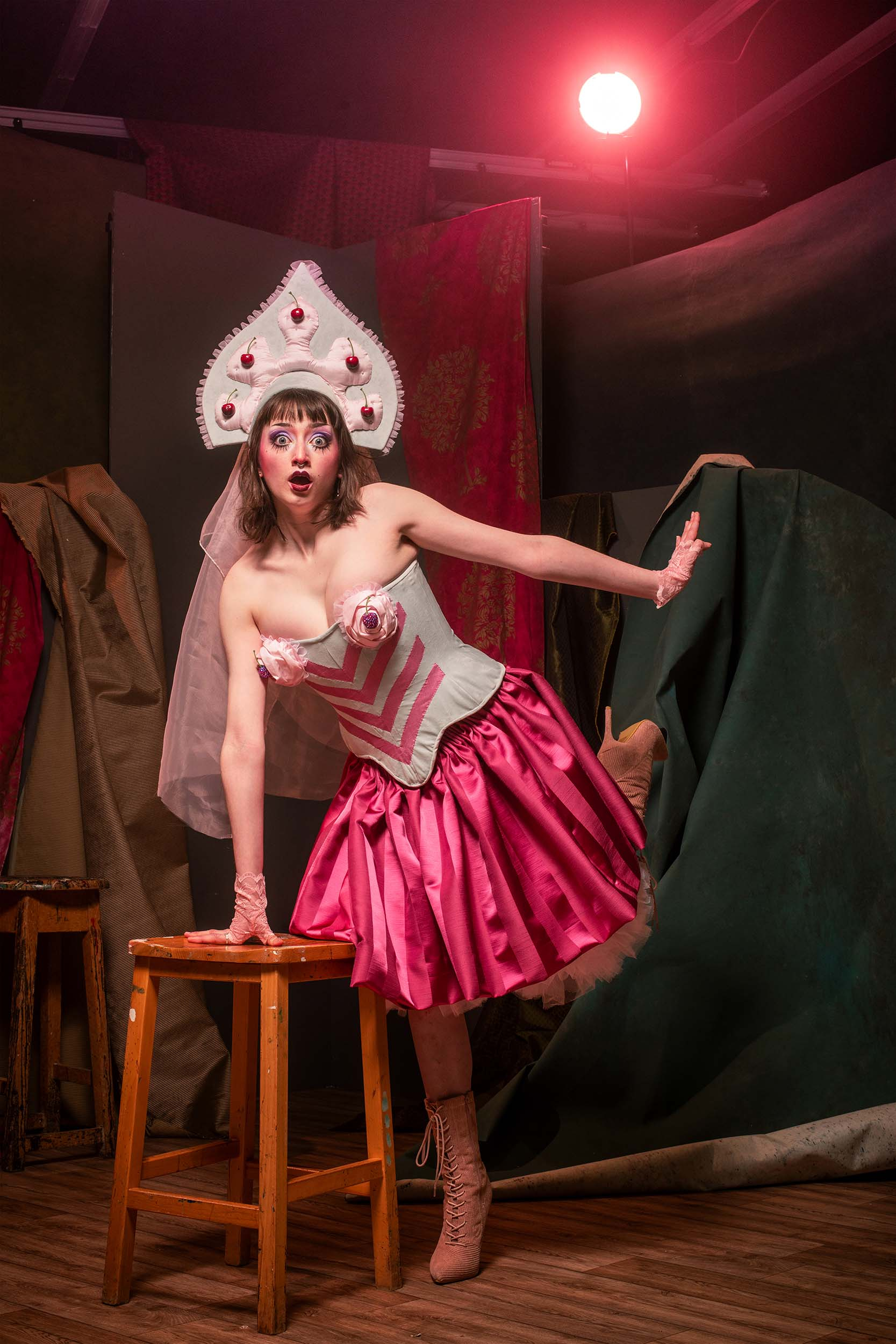 a performer in a corset, skirt and bloomers leans on a chair, looking directly into the camera 