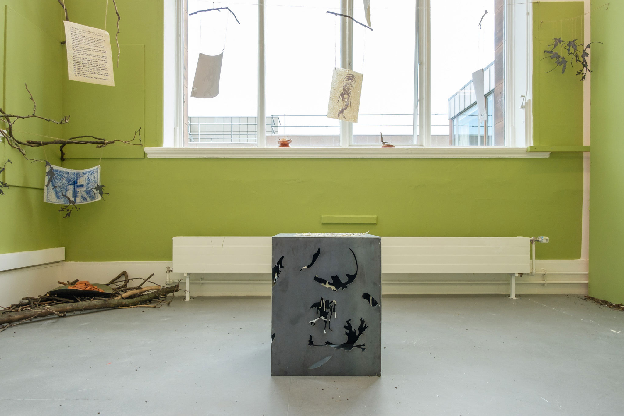 Installation with metal structure in the centre, and handmade paper hanging from branches across the space. metal plate in the back left corner under two prints.