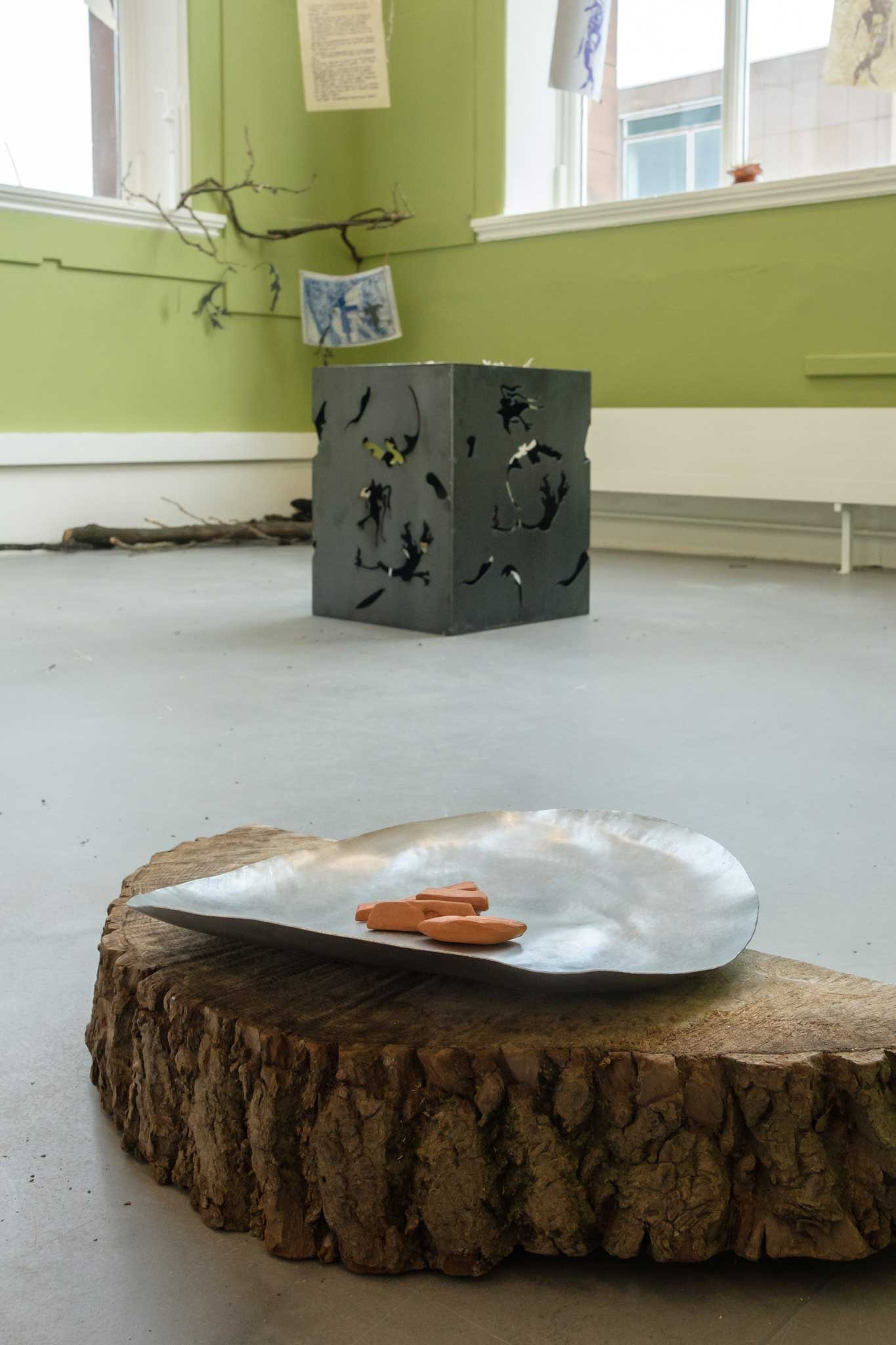 angled view of installation, log with metal tray containing ceramic offerings in the forefront, metal structure and branches in the background. 