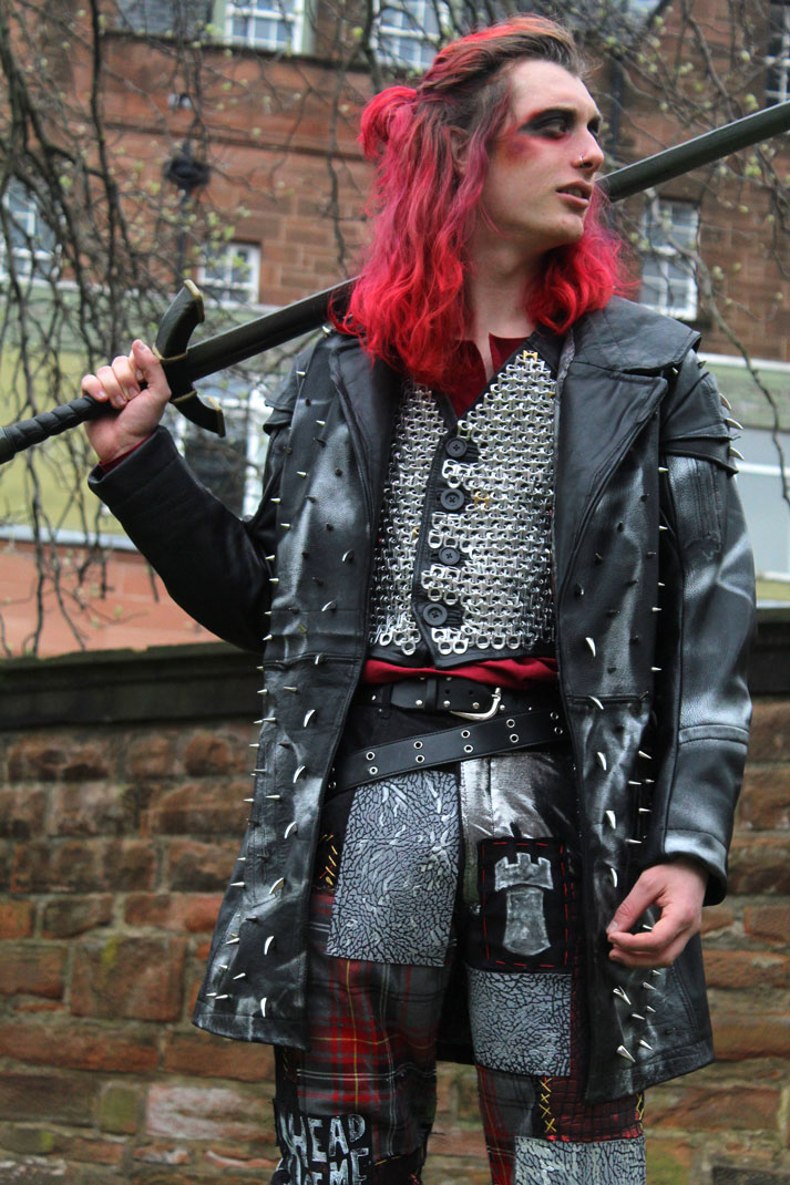 Costume designed based on punk subculture and high fantasy pirates, featuring a tunic, waistcoat covered in recycled chainmail, patched up jeans, and studded leather coat. Performer holds a sword and looks off into the distance.