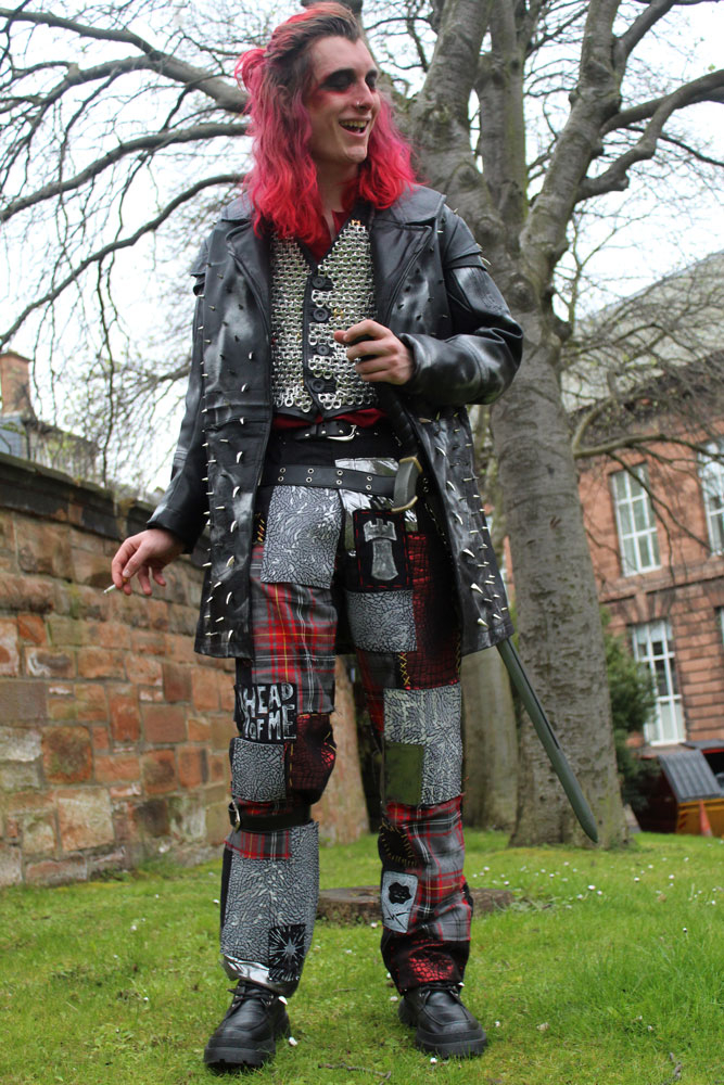 Costume designed based on punk subculture and high fantasy pirates, featuring a tunic, waistcoat covered in recycled chainmail, patched up jeans, and studded leather coat. Performer laughs while holding a cigarette.