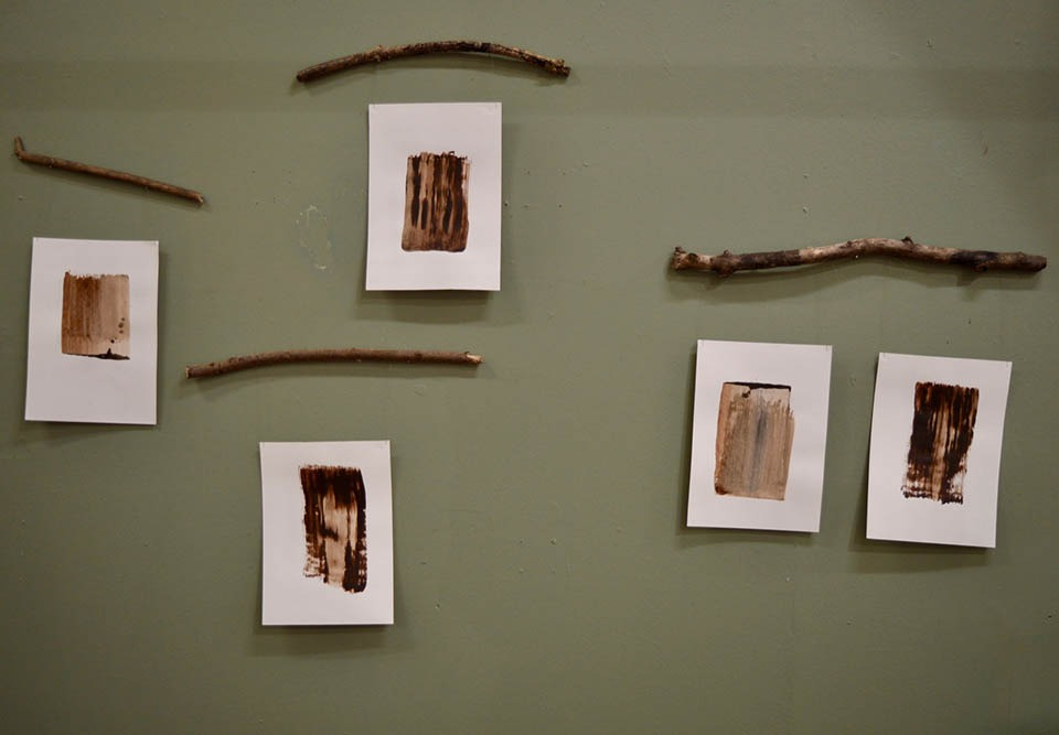A series of single thick brown brushstrokes on white paper, displayed on a green wall and suspended from branches
