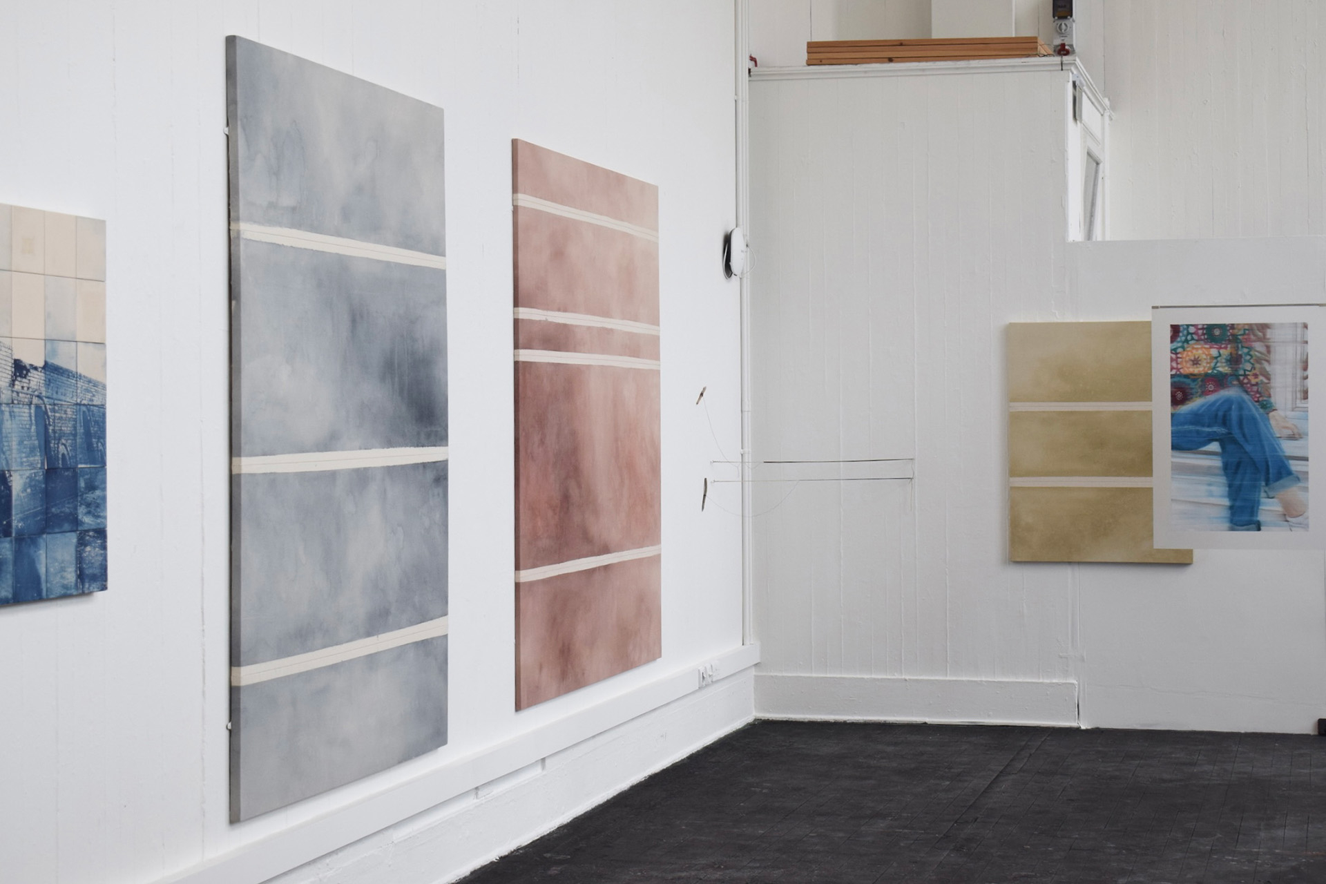 three paintings on a wall. the first one is blue with pale cream stripes, the second is the same but red brown, the third the same with beige-yellow
