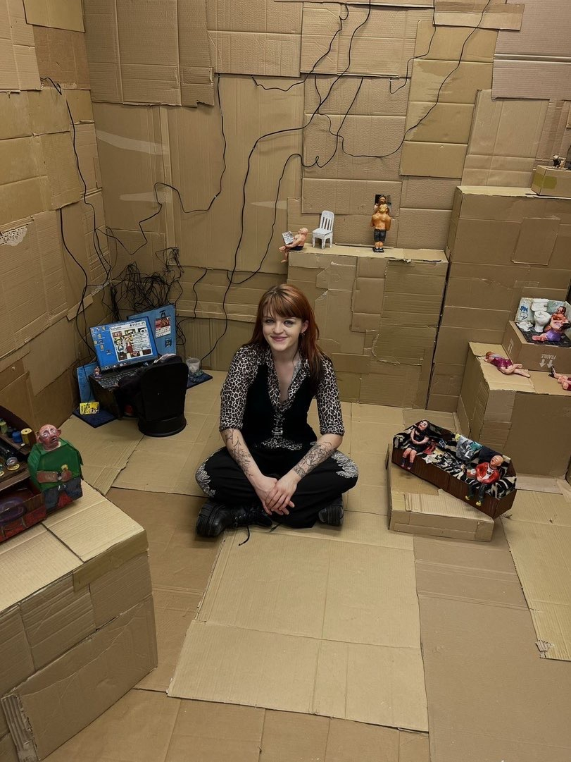 Image of artist within degree show space that is constructed of cardboard. Sculptures placed within this constructed space.