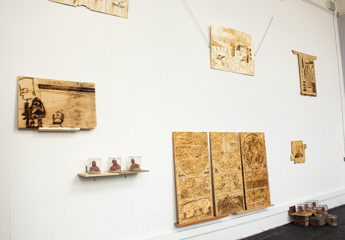 A series of laser engraved wooden panels mounted on a white wall. Accompanying the panels are a series of concrete heads in glass jars.