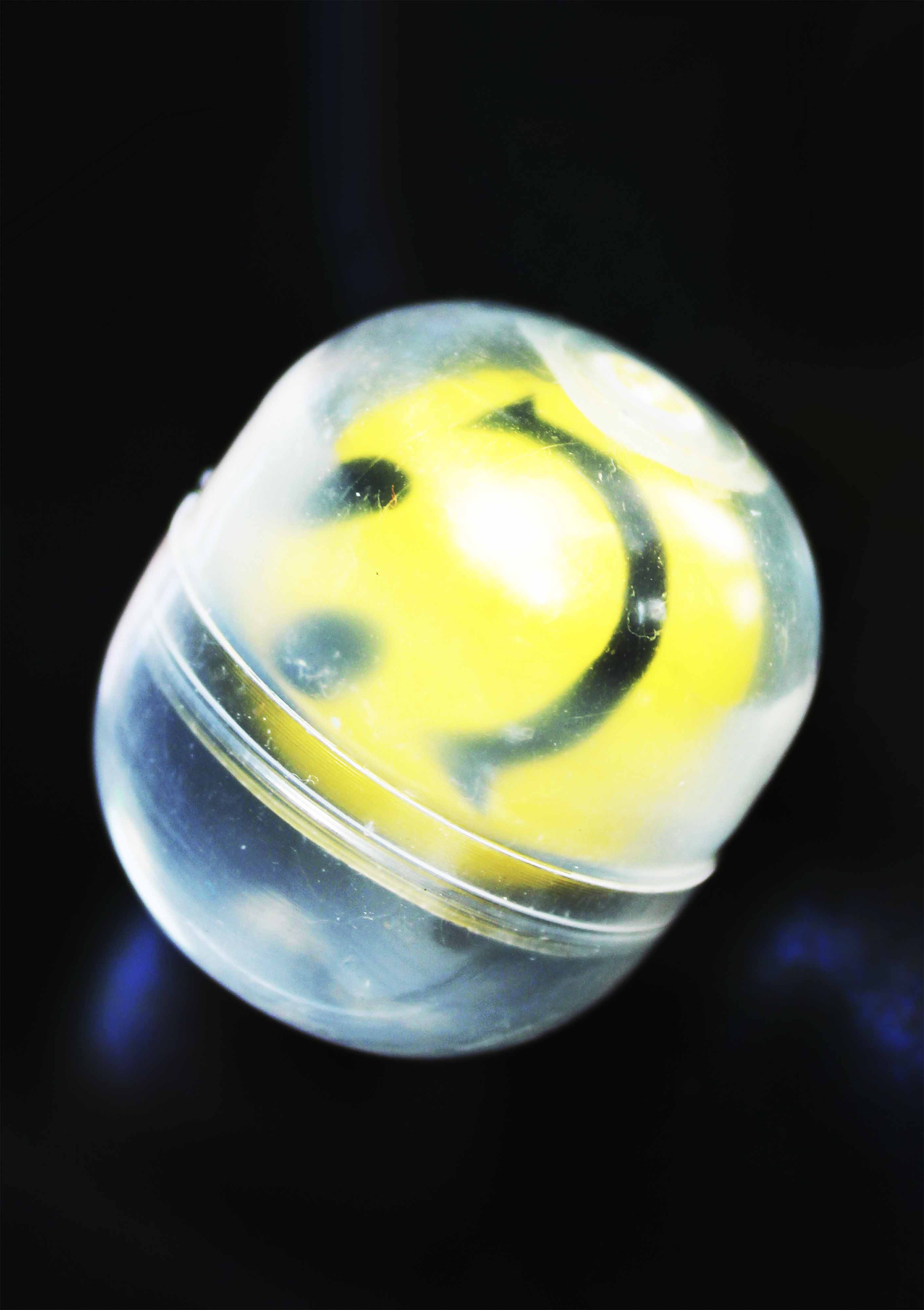 Image captures a yellow smiley face bouncy ball contained in a plastic capsule 
